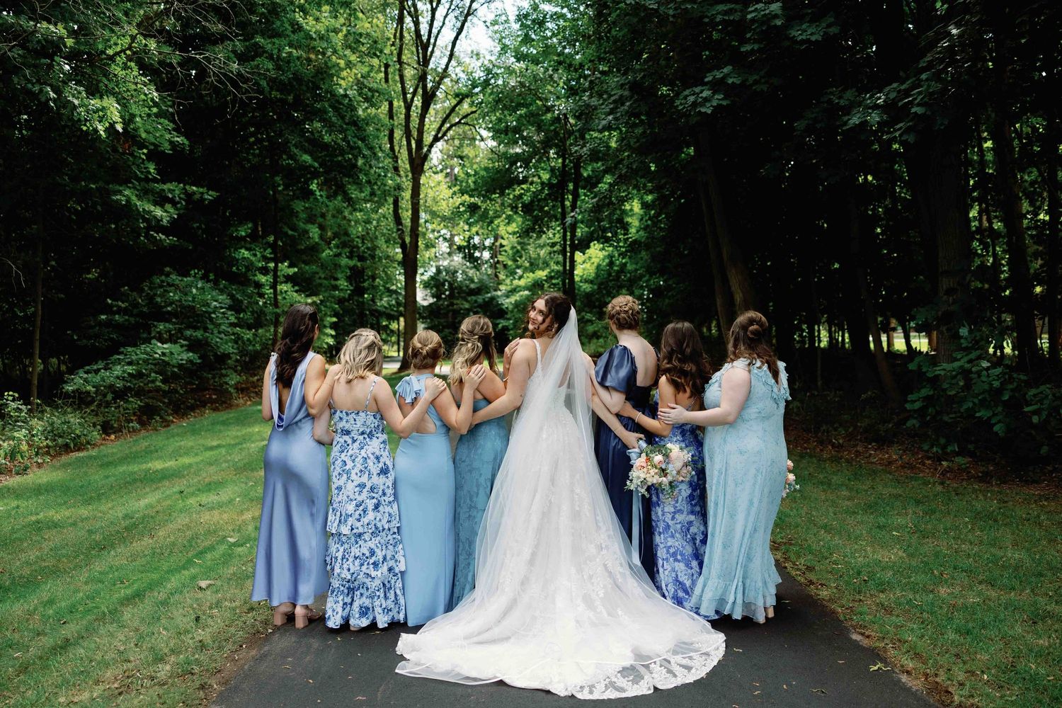 Bridesmaids in light blue dresses pose with the bride in a white gown on a grassy outdoor path.