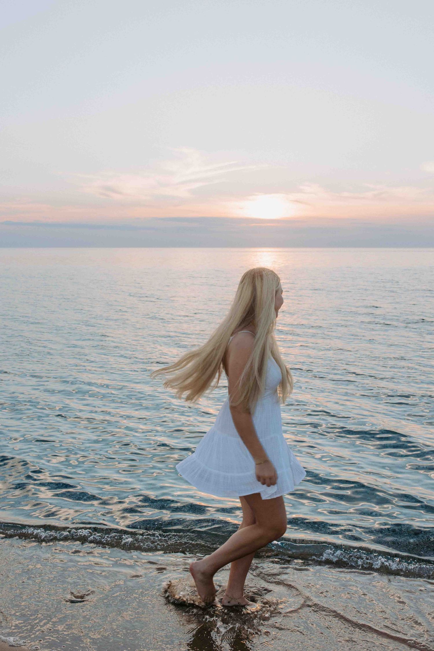 A person in a white dress walks along a scenic beach shoreline at sunset with calm ocean waters.