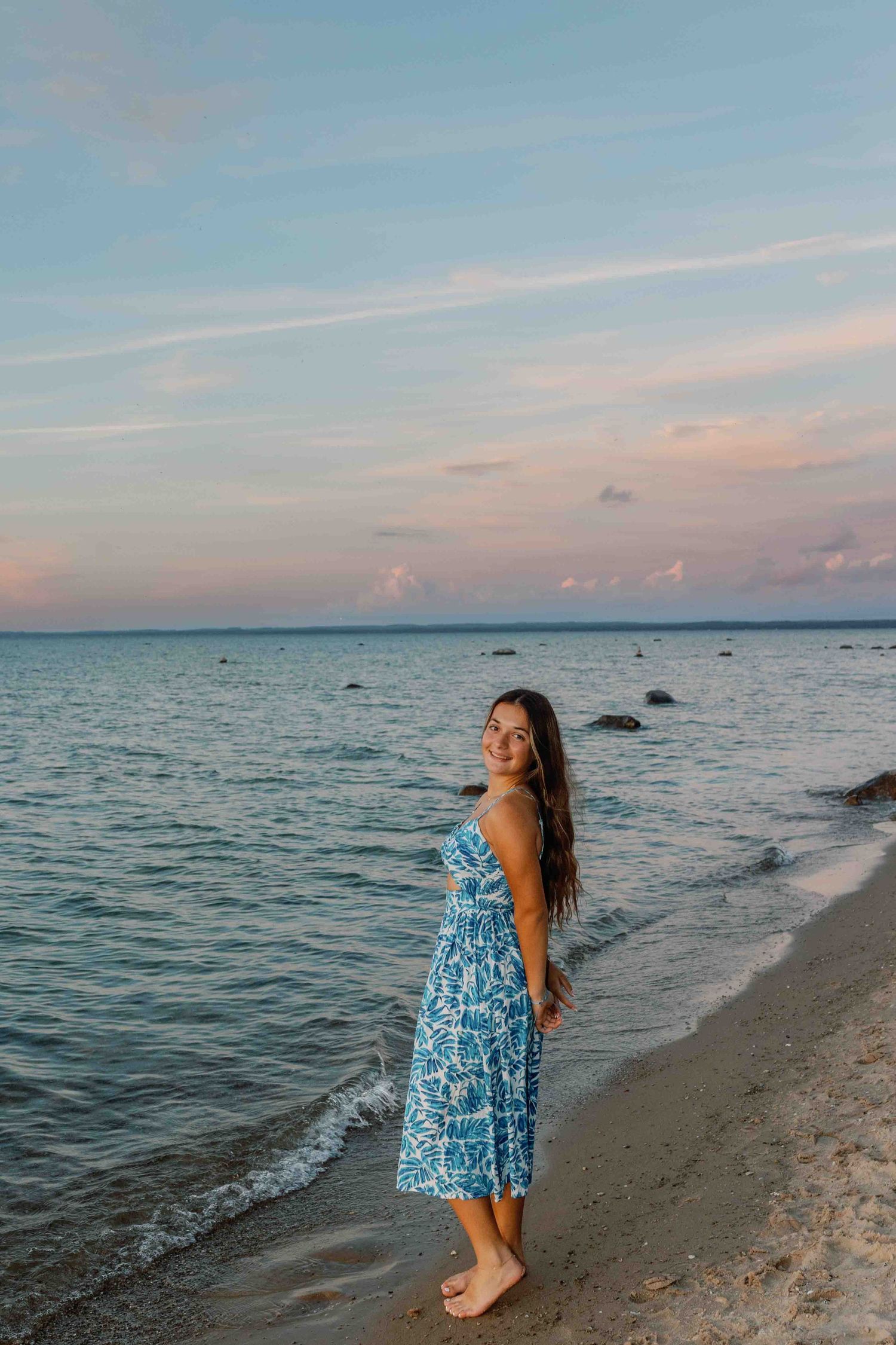A blue and white patterned sundress flows in the breeze as someone poses along a serene beach at sunset.