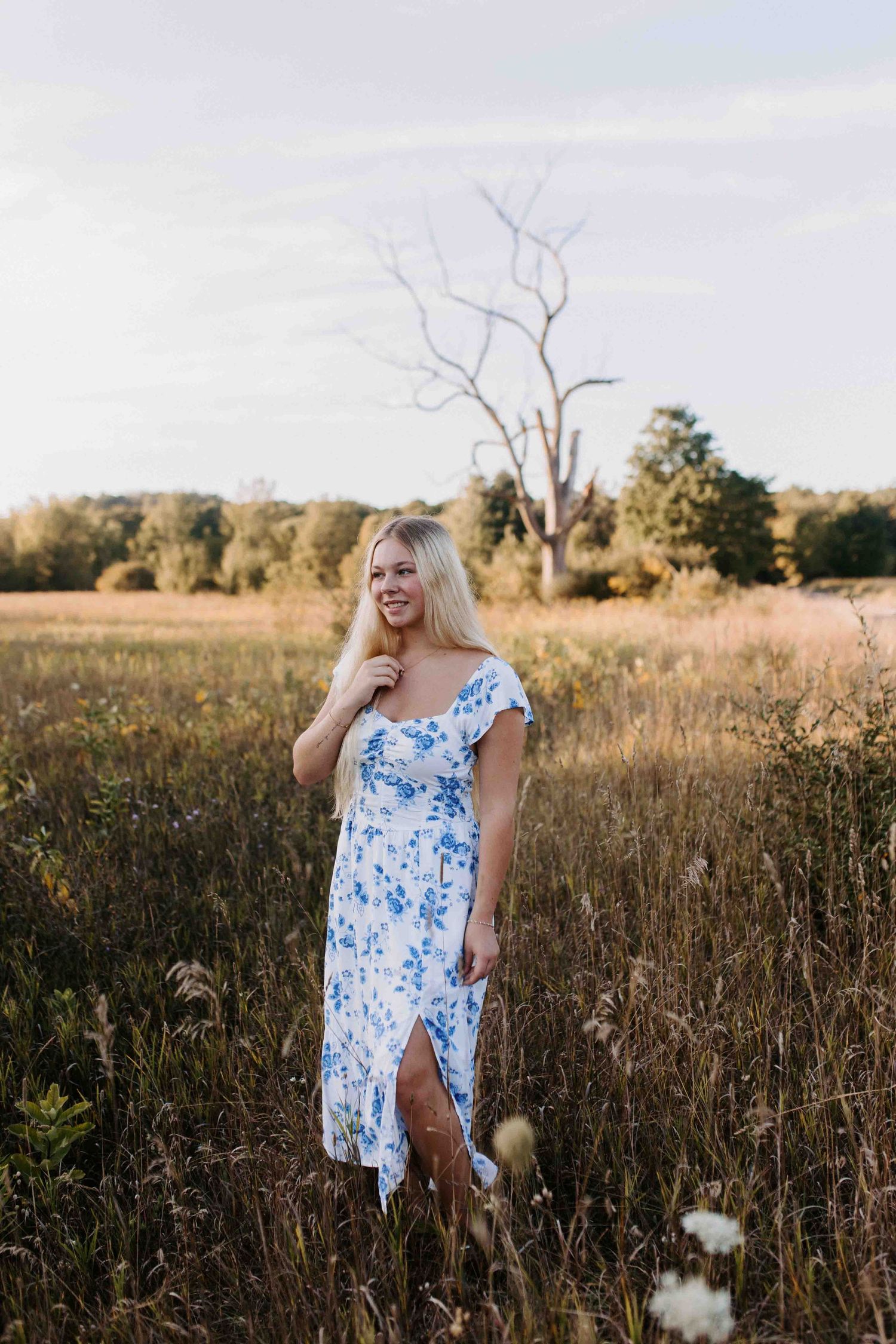 Person in floral dress stands among tall grass in golden evening light.