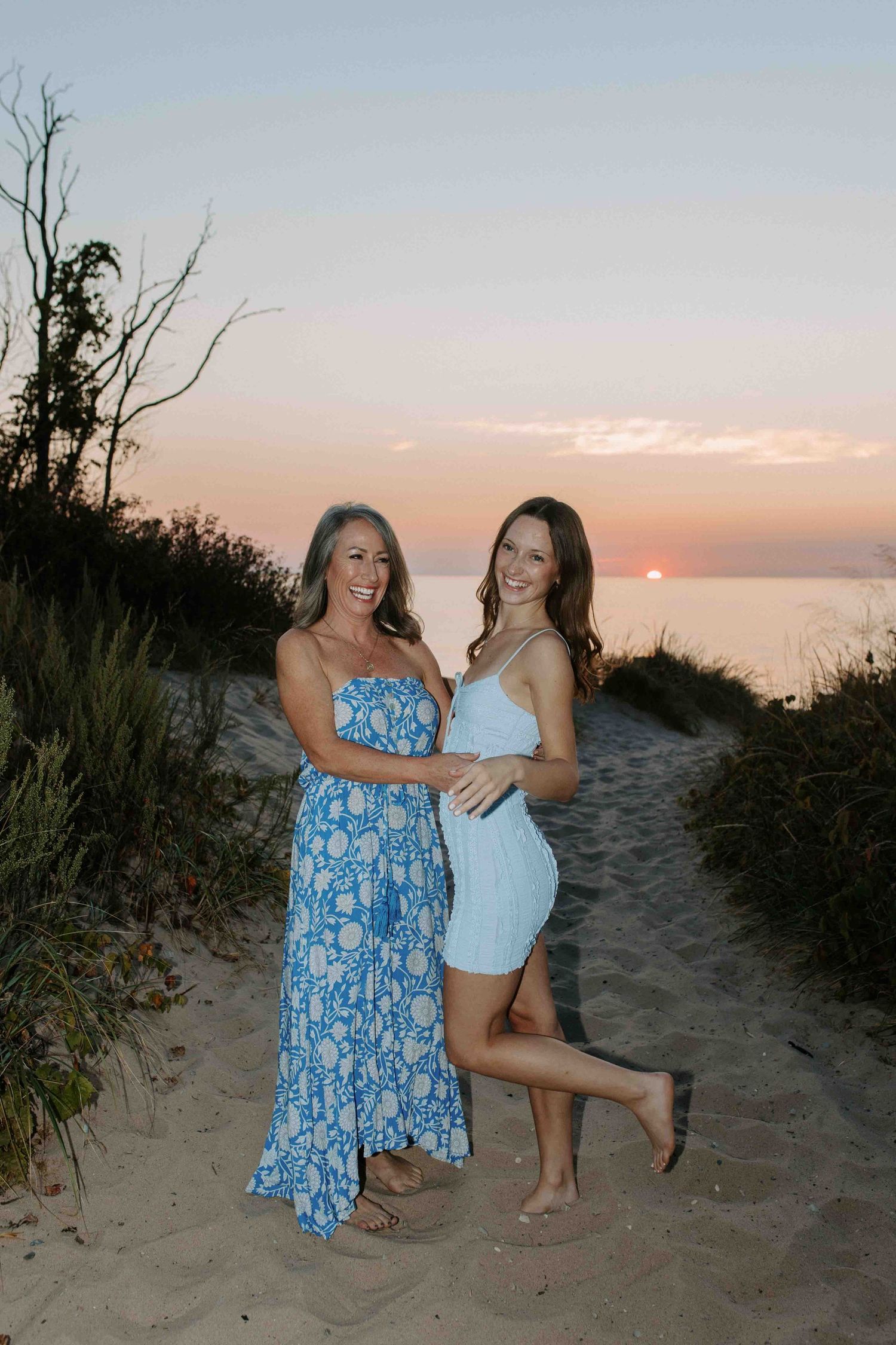 Two people in summer dresses pose playfully on a sandy beach path during sunset.