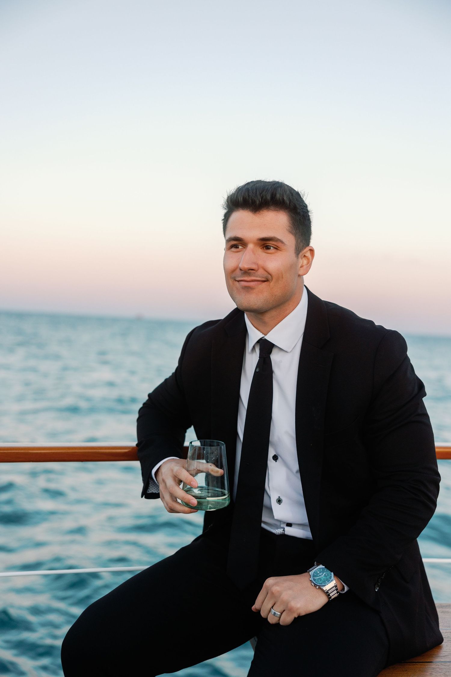 Groom in black tuxedo relaxing on boat deck with city skyline in background.