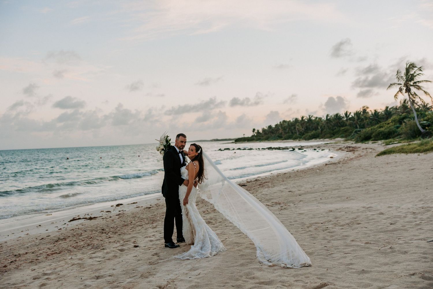 Panoramic beach wedding scene with palm trees and guests gathered along the shoreline.