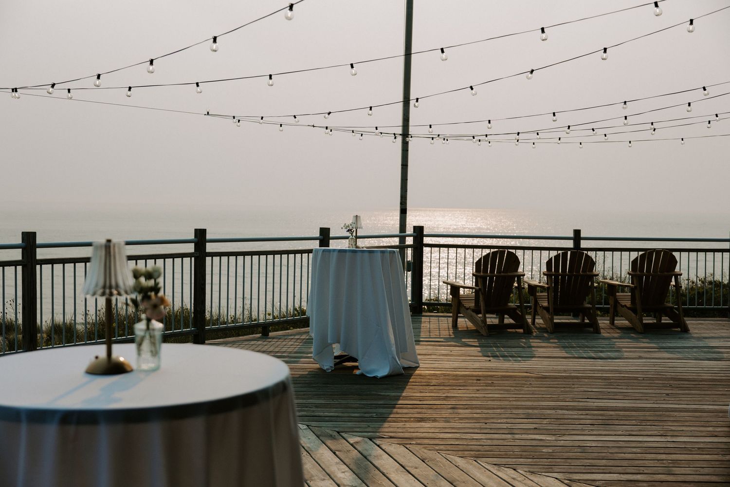 String lights hanging over a wooden deck with ocean views at dusk.