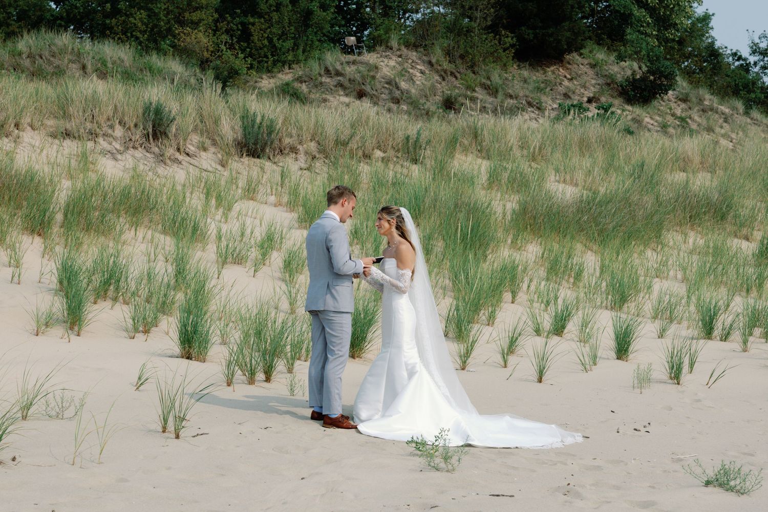 Wedding couple walks along a scenic beach path with grassy dunes on a sunny day.