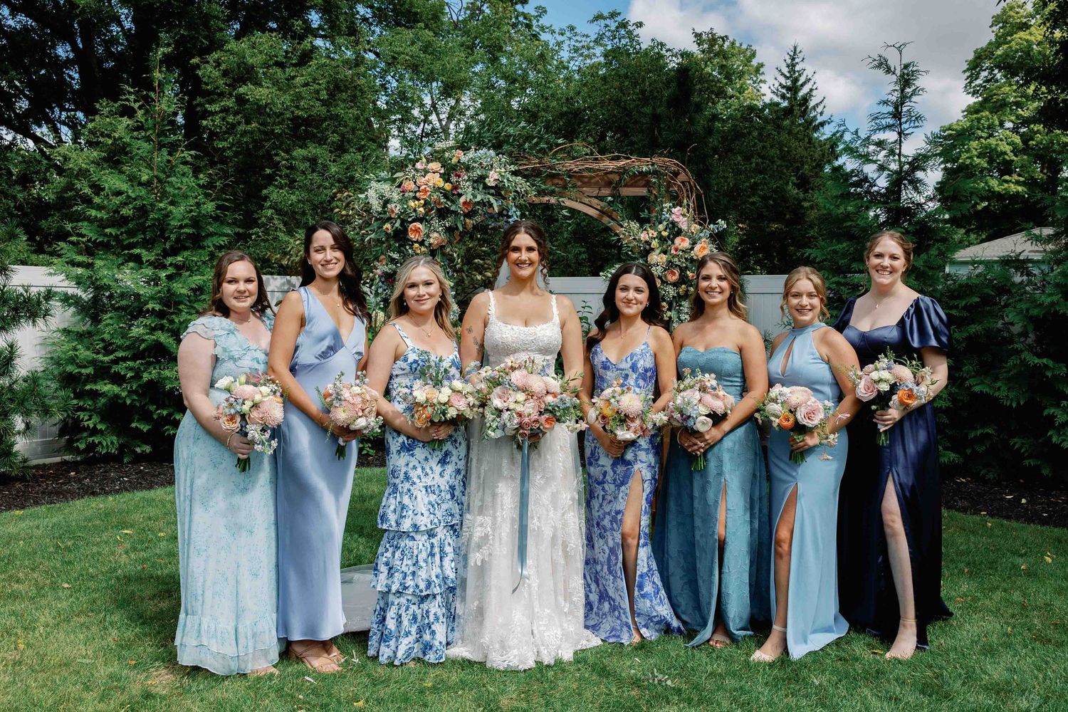 Bridal party in coordinated blue dresses lined up for a group photo on a grassy lawn.