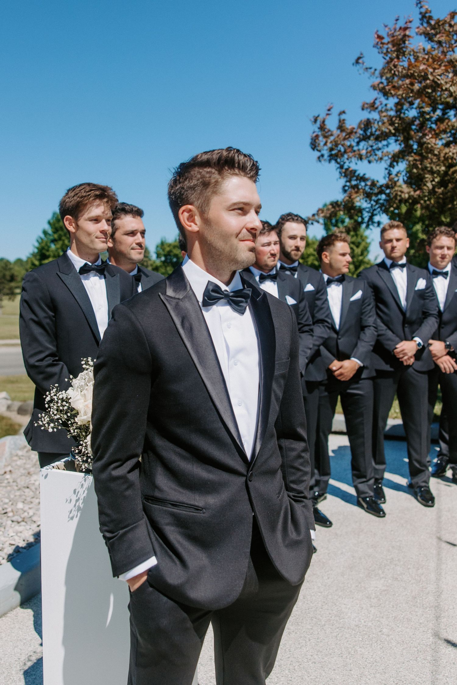 Groomsmen in black tuxedos lined up outdoors on a sunny day.
