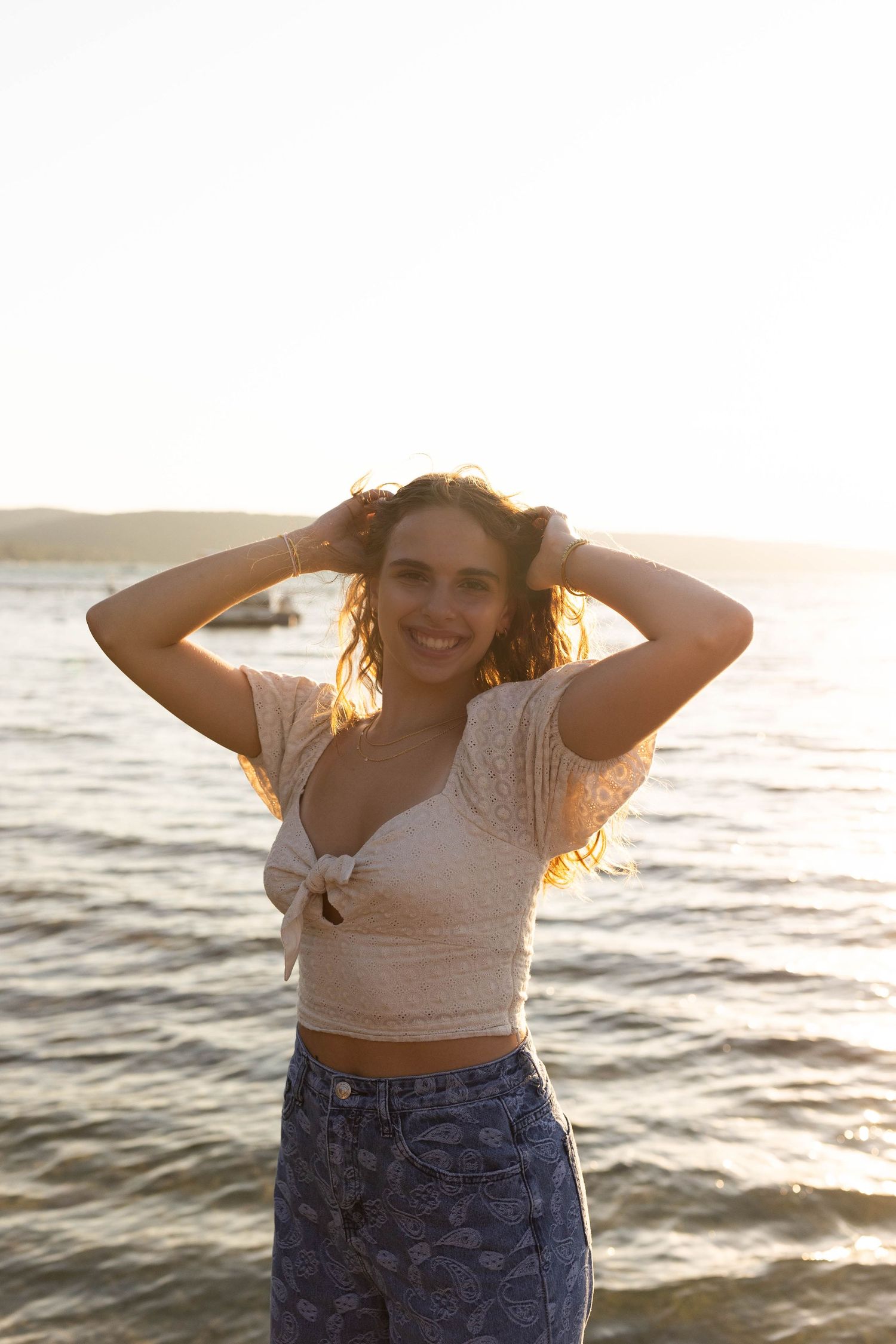 Woman in casual attire poses playfully at beach during golden hour.
