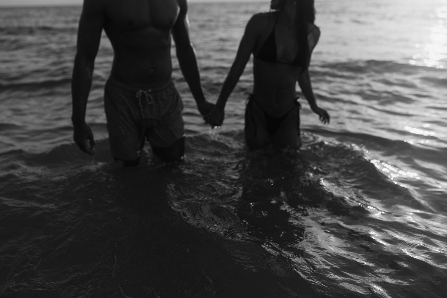 Silhouetted legs stand in shallow ocean water during sunset.