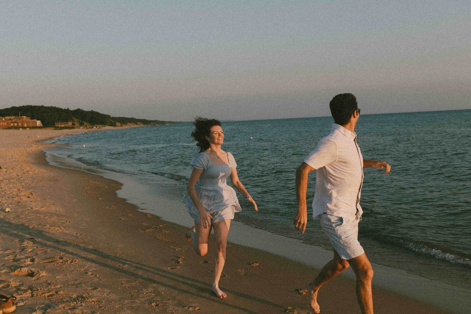 A couple walks playfully along the shoreline during golden hour with a lighthouse visible in the distance.