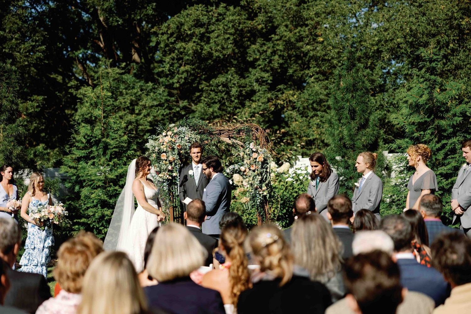 Wedding ceremony guests seated in rows outdoors surrounded by green trees.