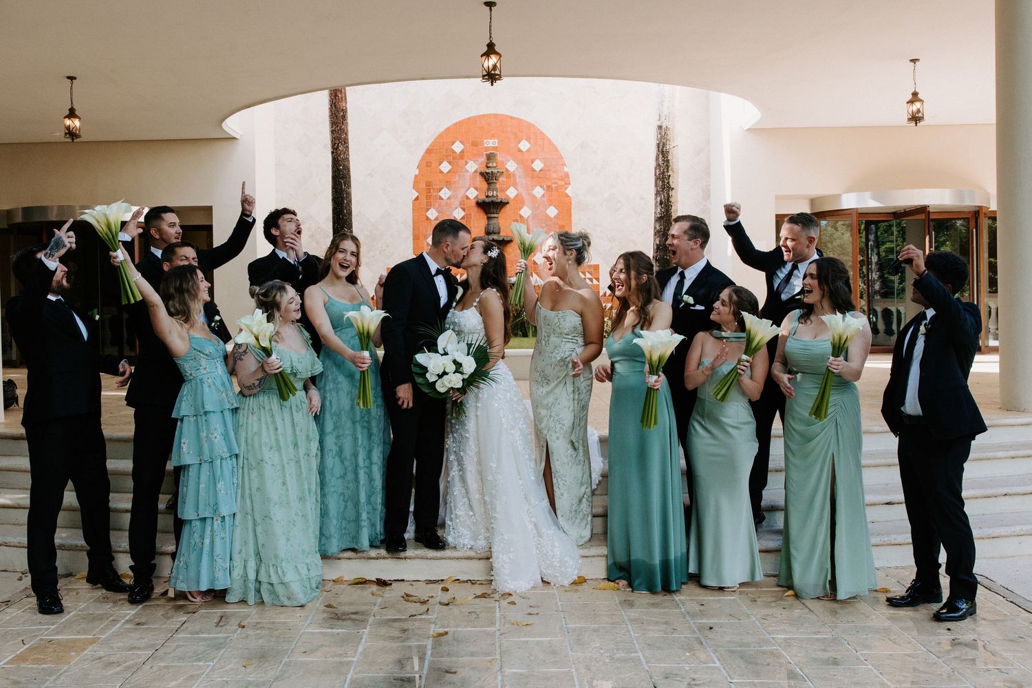 Wedding party in turquoise dresses celebrates with raised arms on outdoor terrace.