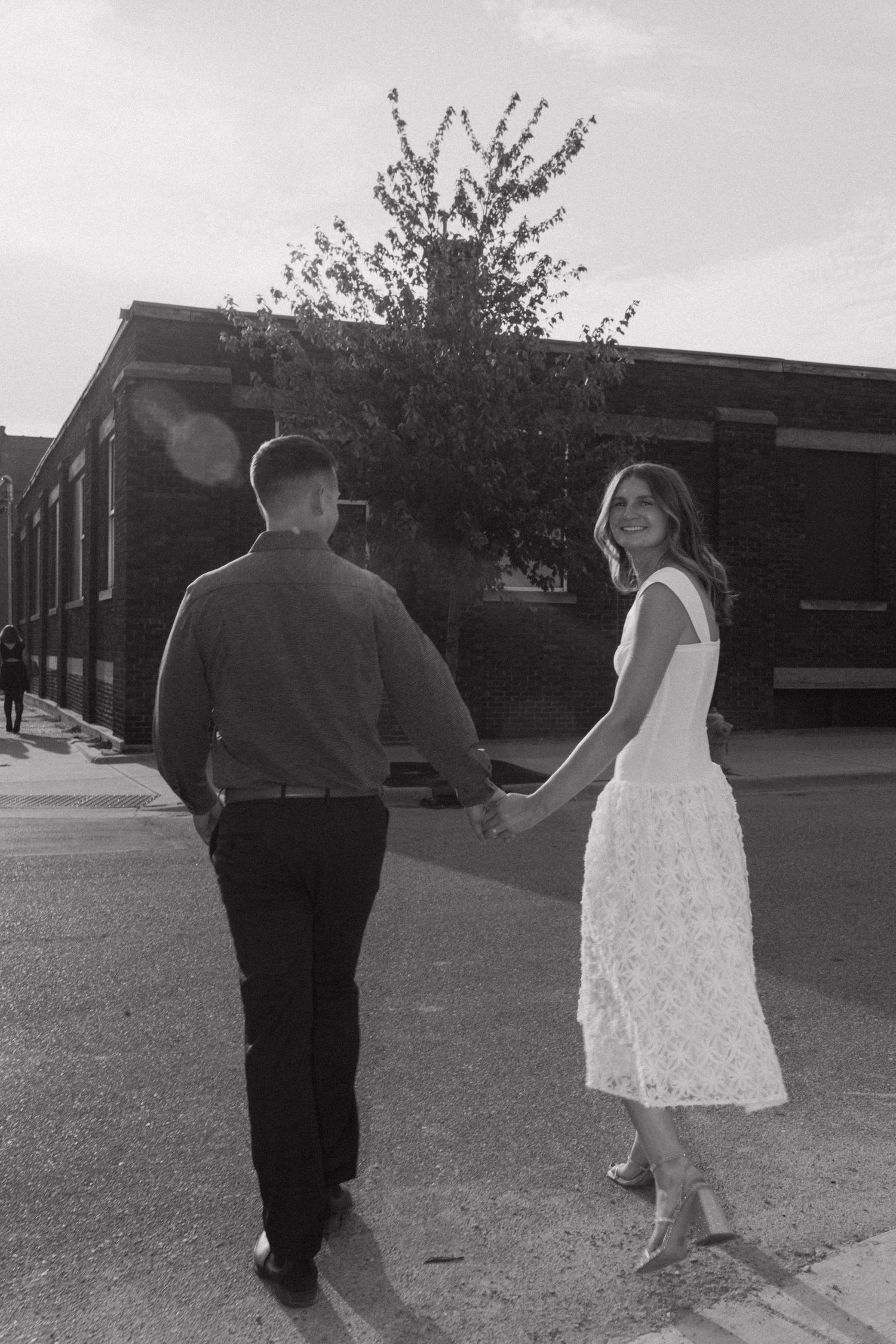 A couple walks through a park in black and white photography with blooming trees in background.