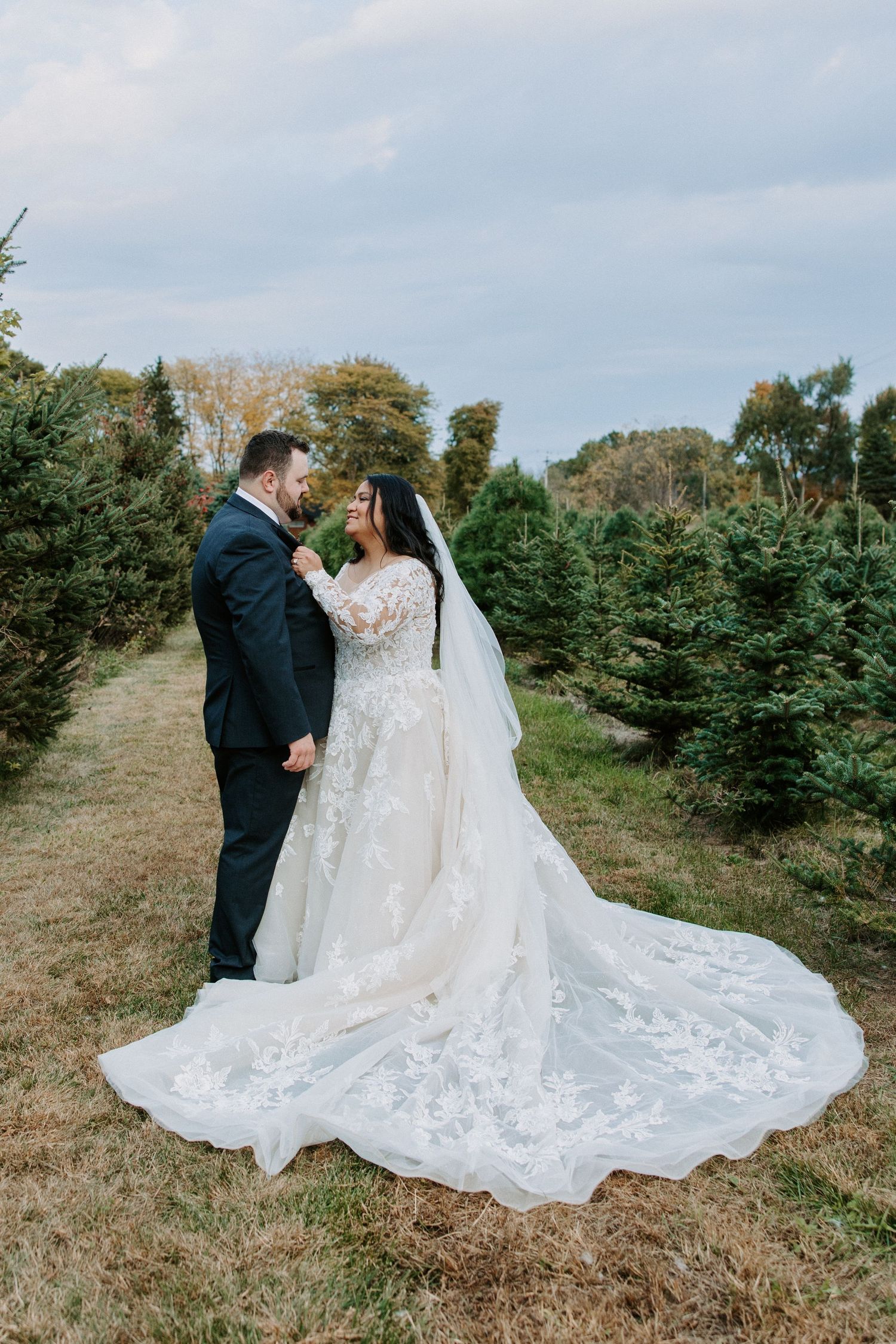 Wedding couple poses romantically among Christmas trees in flowing lace gown.