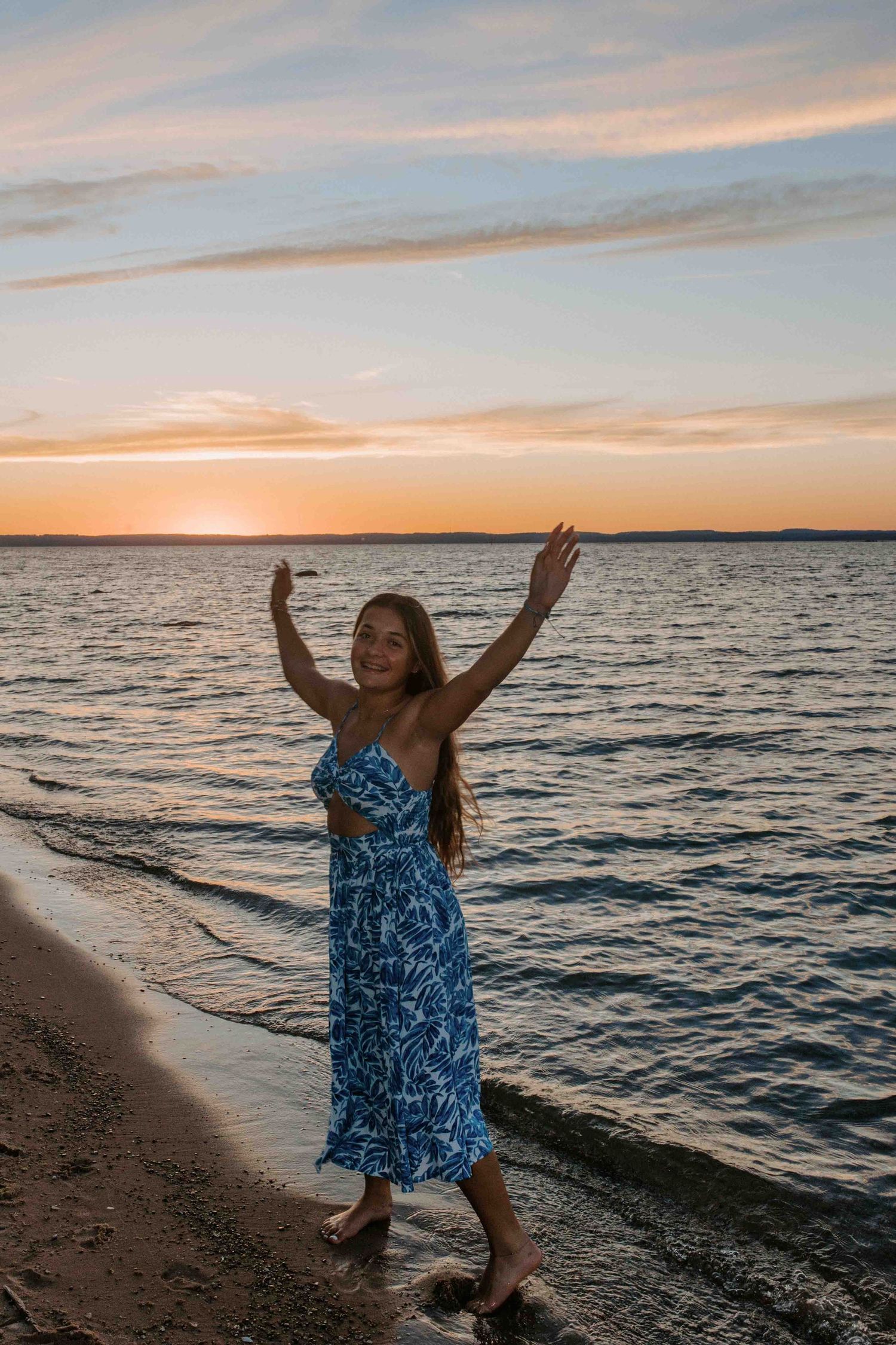 Figure in blue sundress raises arms joyfully on beach during golden hour.