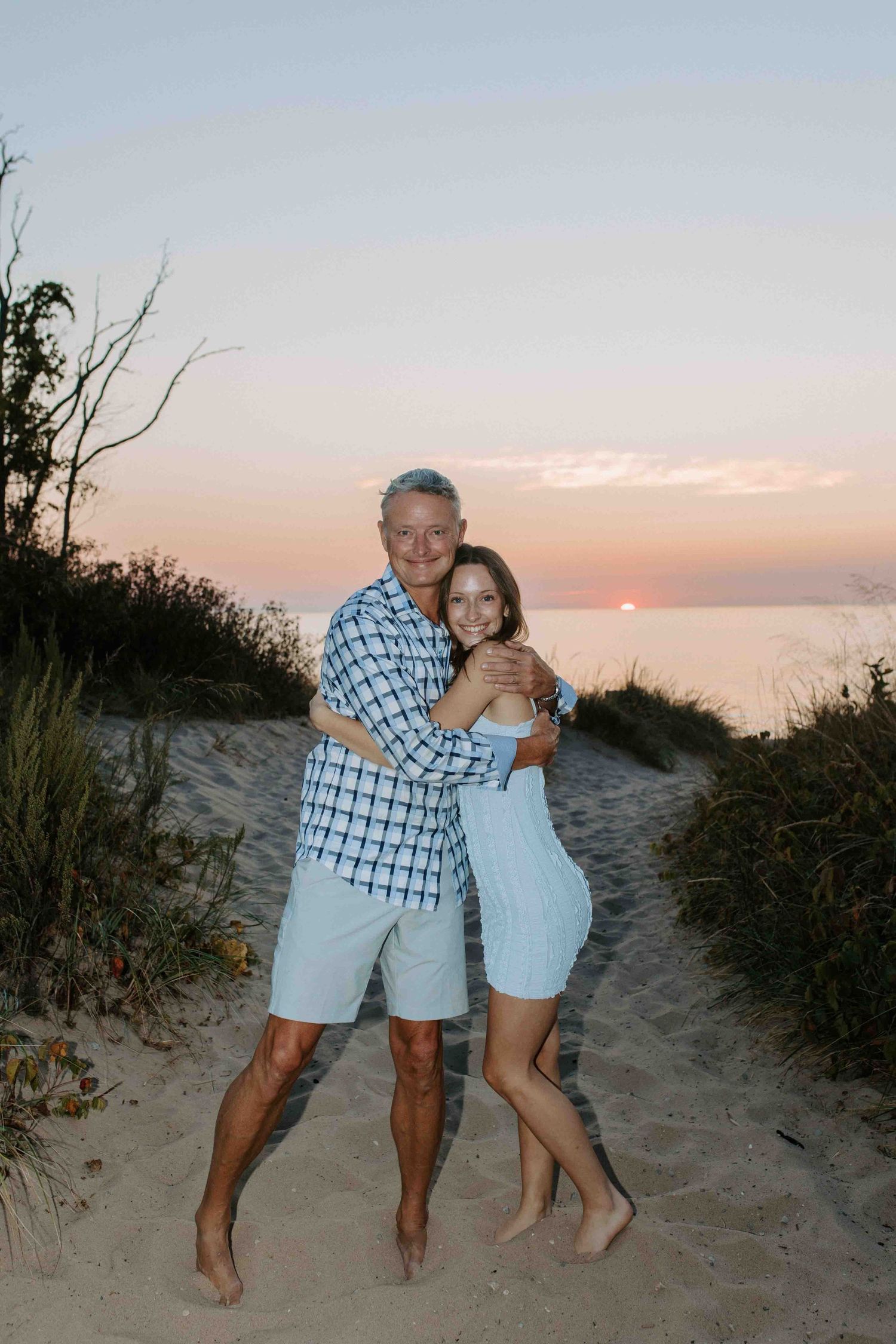 Two people embrace while standing barefoot on a sandy beach trail at sunset.
