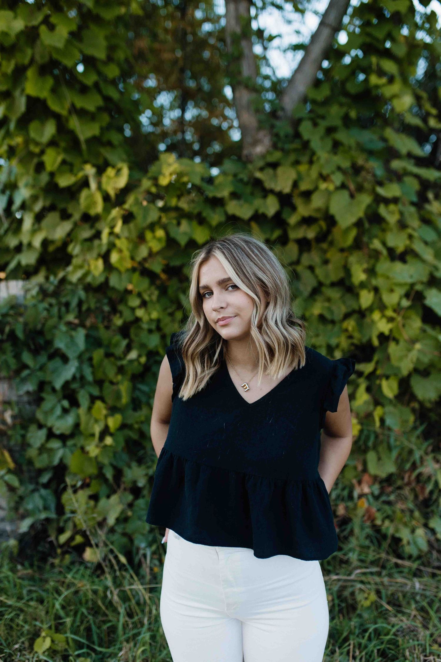Casual outdoor portrait against lush green foliage wearing black top and white pants.
