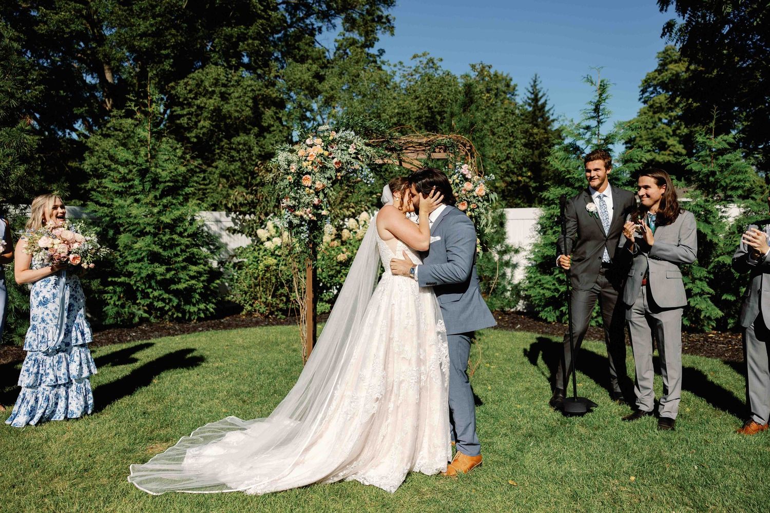 Wedding ceremony guests gather on white chairs on a grassy lawn under blue skies.