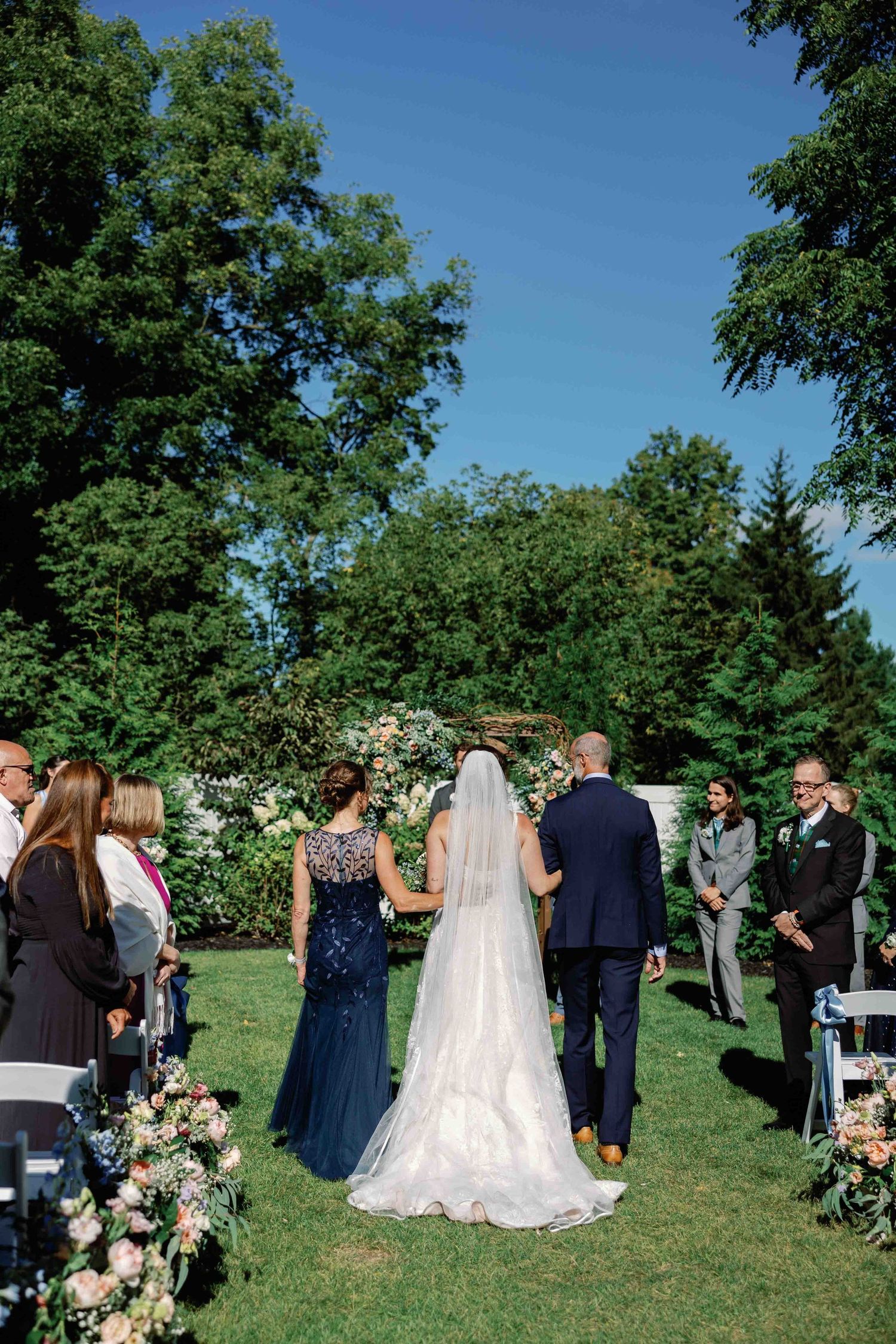 Wedding ceremony taking place on a grassy lawn with floral decorations under blue skies.