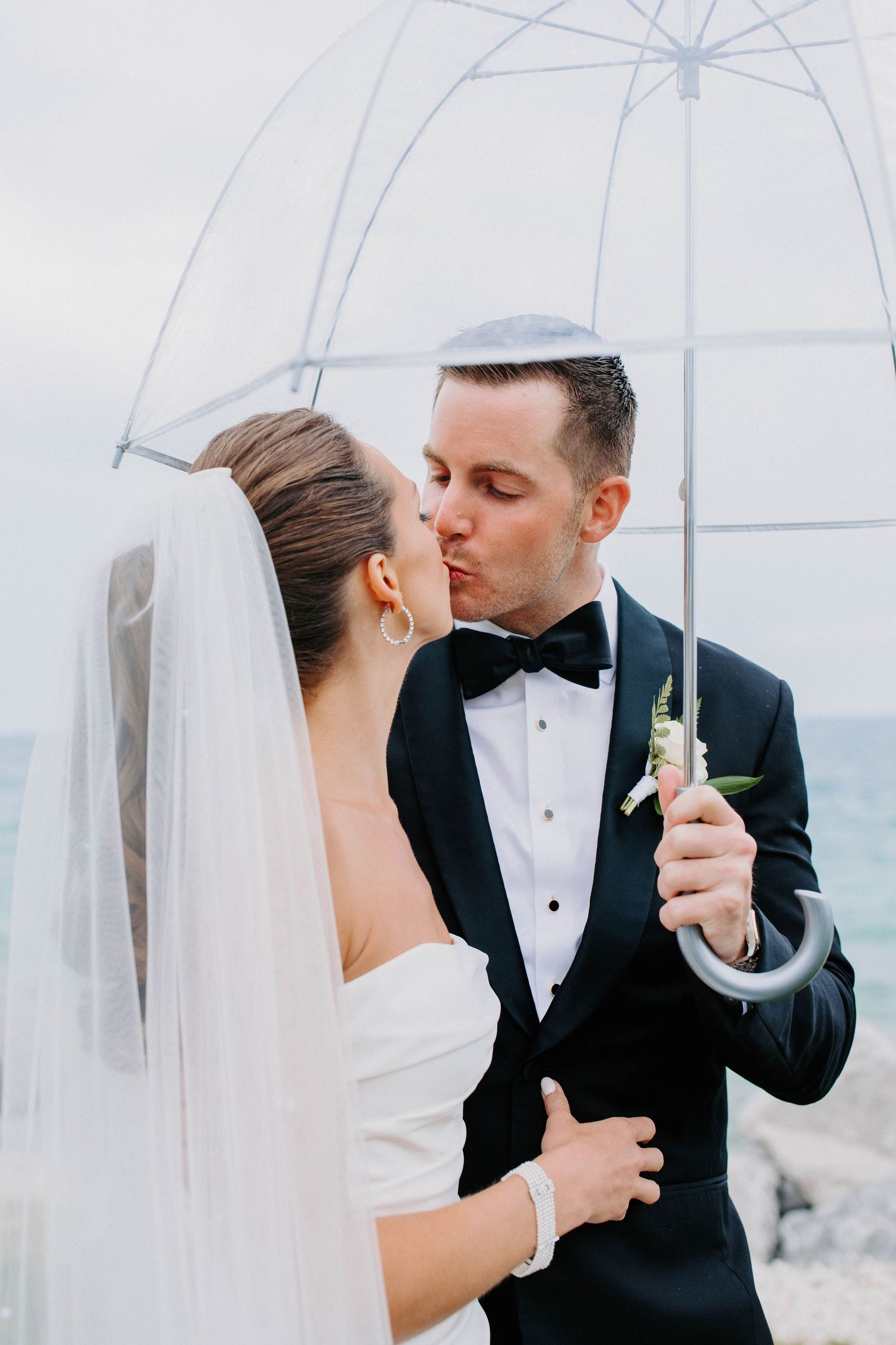 A romantic wedding portrait of a couple embracing under a clear umbrella with ocean views.