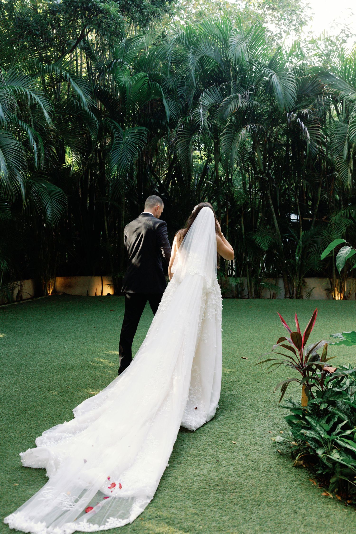 Wedding couple walking on grass with long white train trailing behind in tropical garden setting.