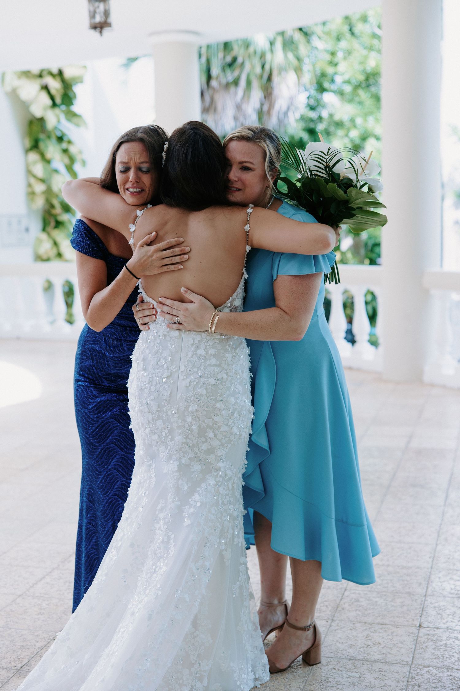 Emotional group hug between bride in white dress and bridesmaids in blue on a columned porch.
