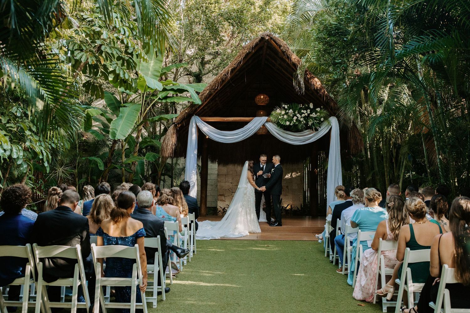 Outdoor wedding ceremony setup with white chairs and tropical garden backdrop.