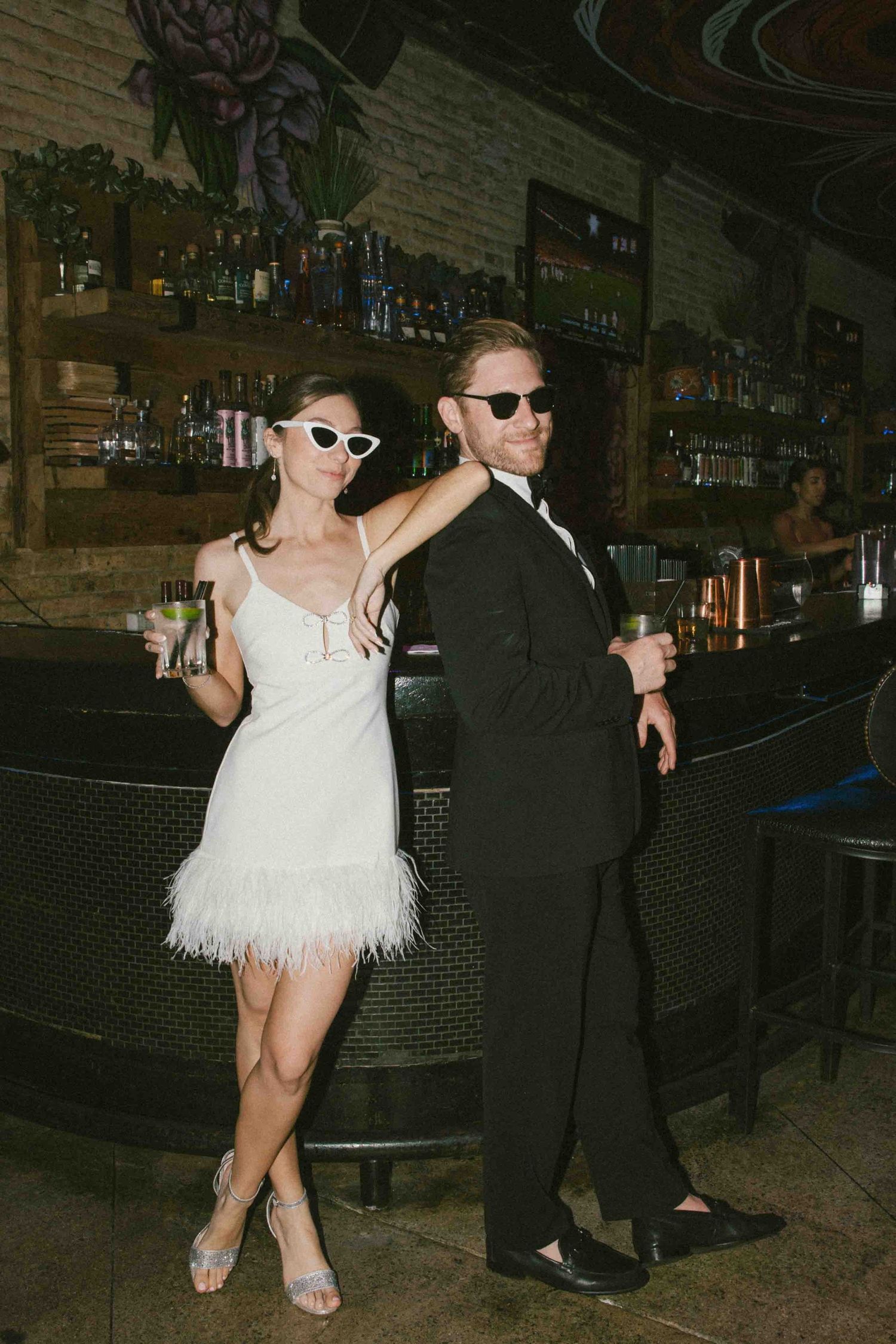 A couple poses playfully at a bar with exposed brick walls and neon signage.