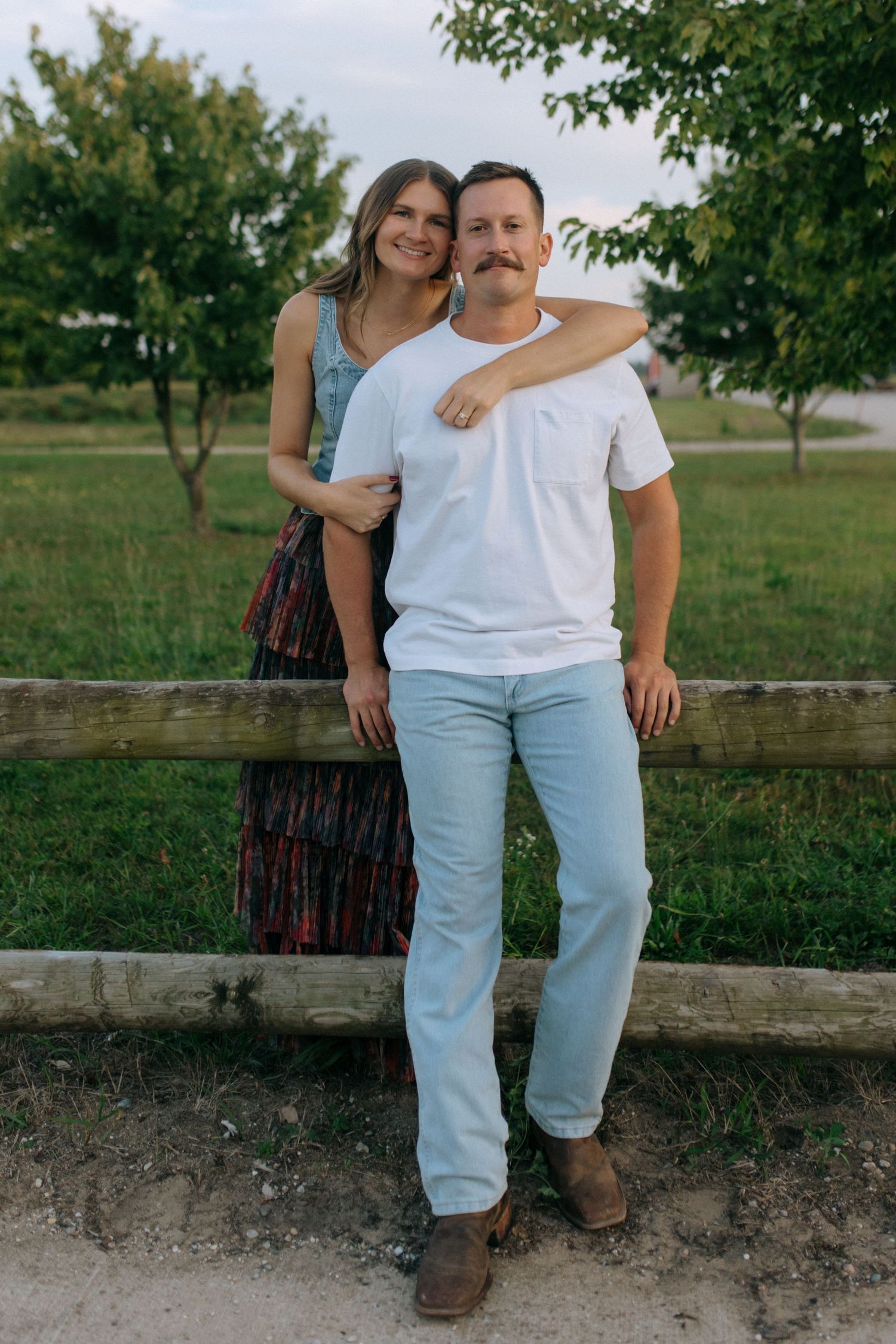 A couple sits together on a rustic wooden fence in a countryside setting.
