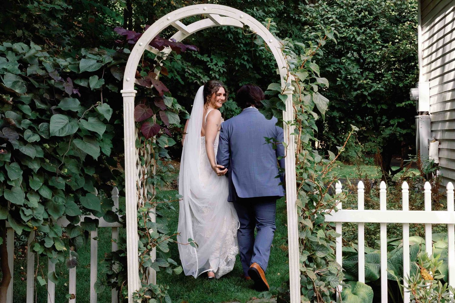 Wedding couple walks through a garden path with white picket fence and green foliage.