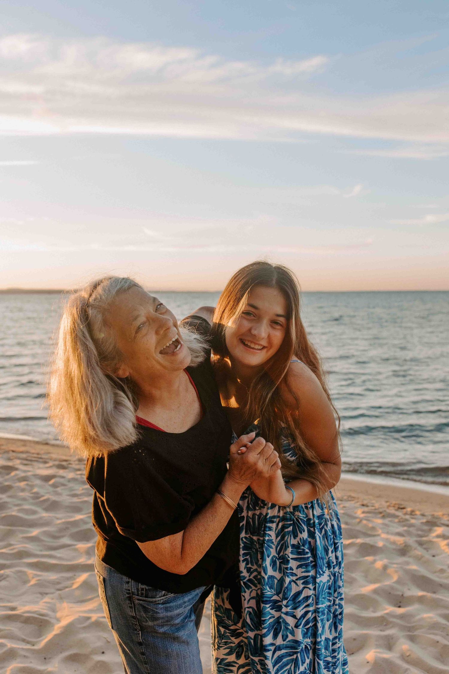Two people laugh together while standing on a beach during golden hour.