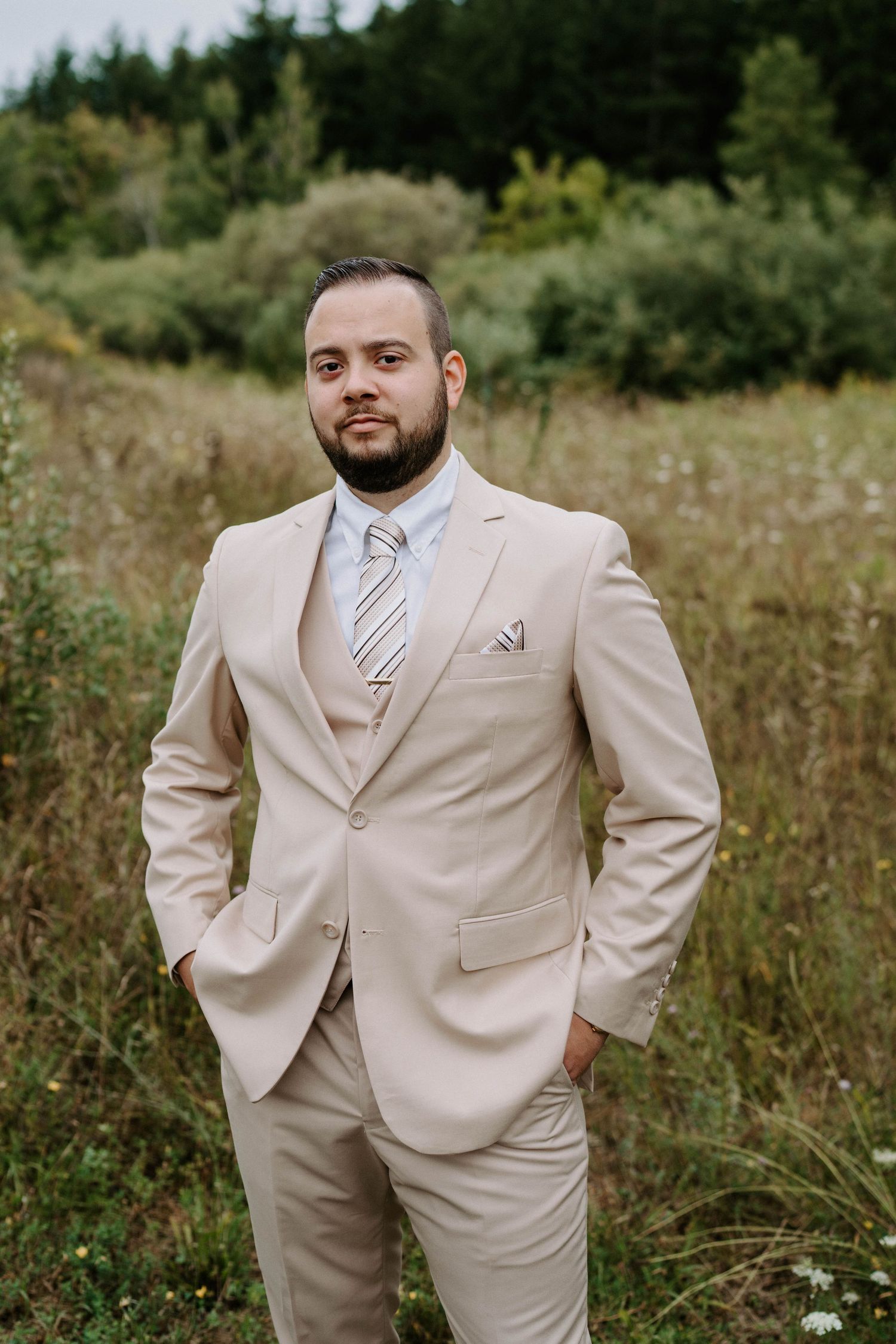 Groom poses confidently in beige suit against natural wildflower backdrop.