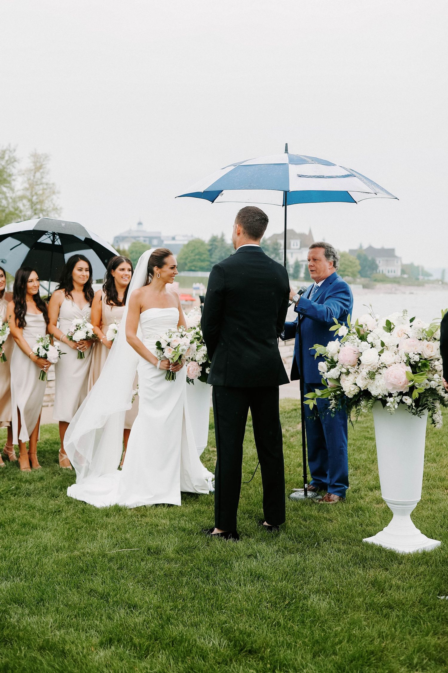 Wedding party stands on grass with umbrellas and white floral arrangements during an overcast outdoor ceremony.