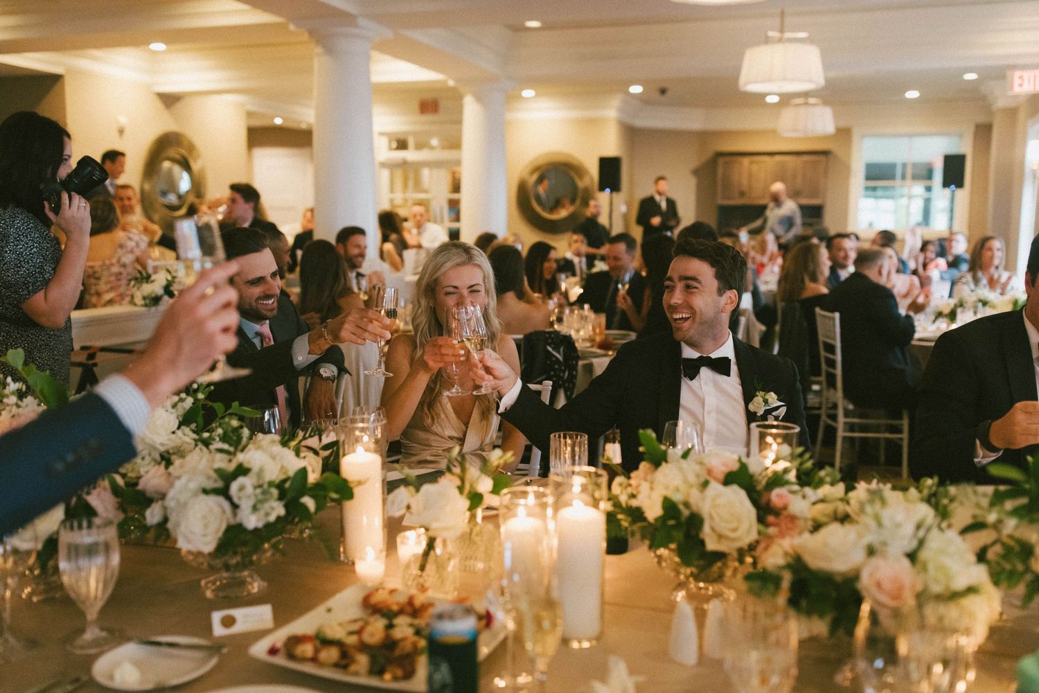 Warm-toned photo of wedding guests toasting during reception dinner with elegant table decorations.