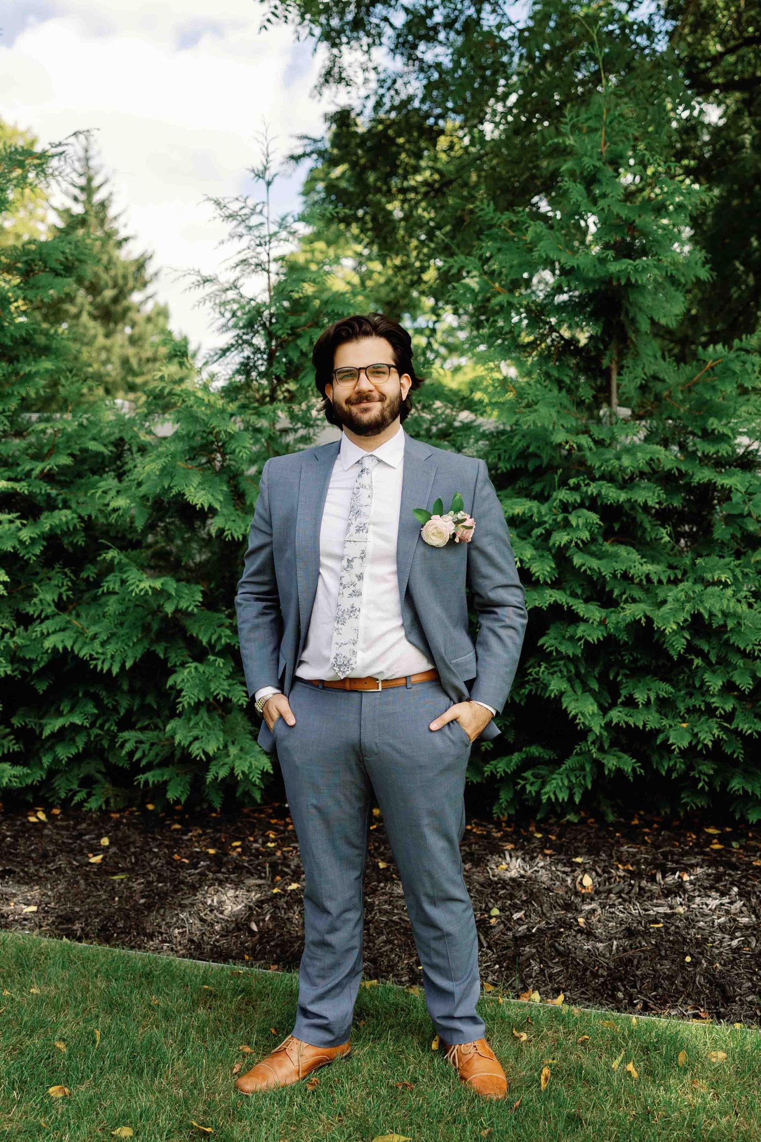 A groom in a gray suit and brown shoes poses against a backdrop of evergreen trees.