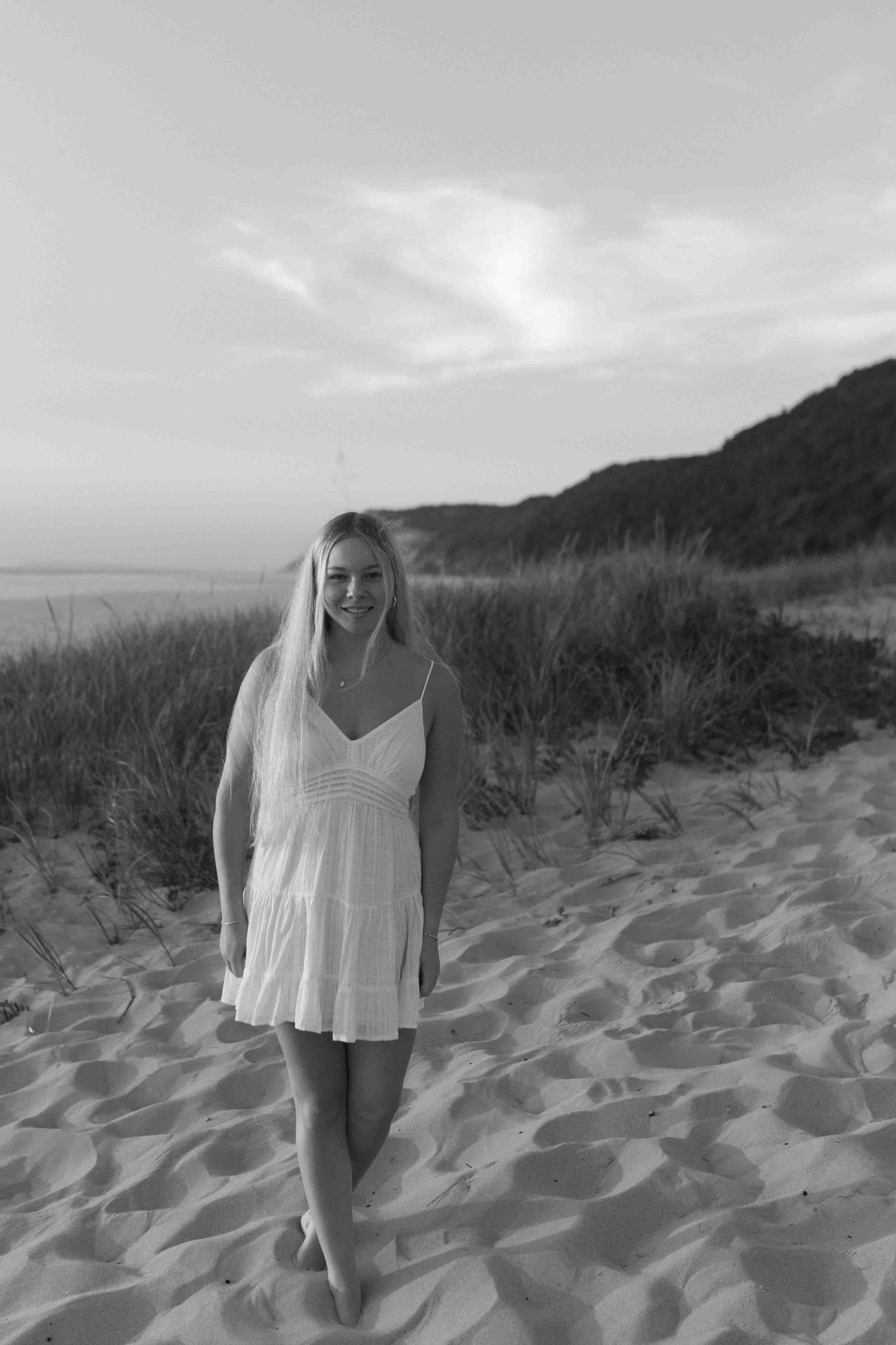 Black and white photo of someone in a white dress standing on beach sand dunes.
