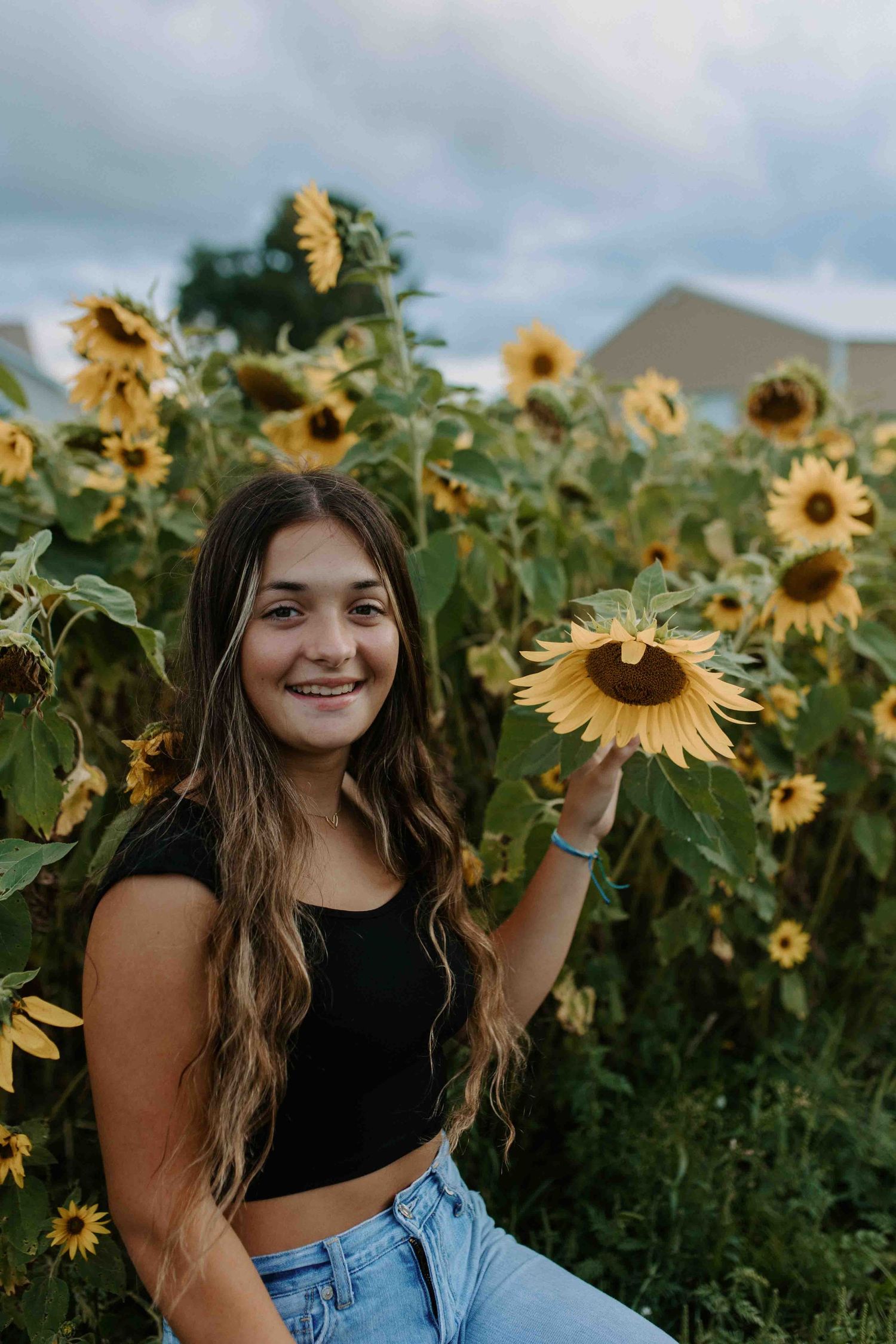 Person in black top and jeans poses joyfully in a field of bright yellow sunflowers.