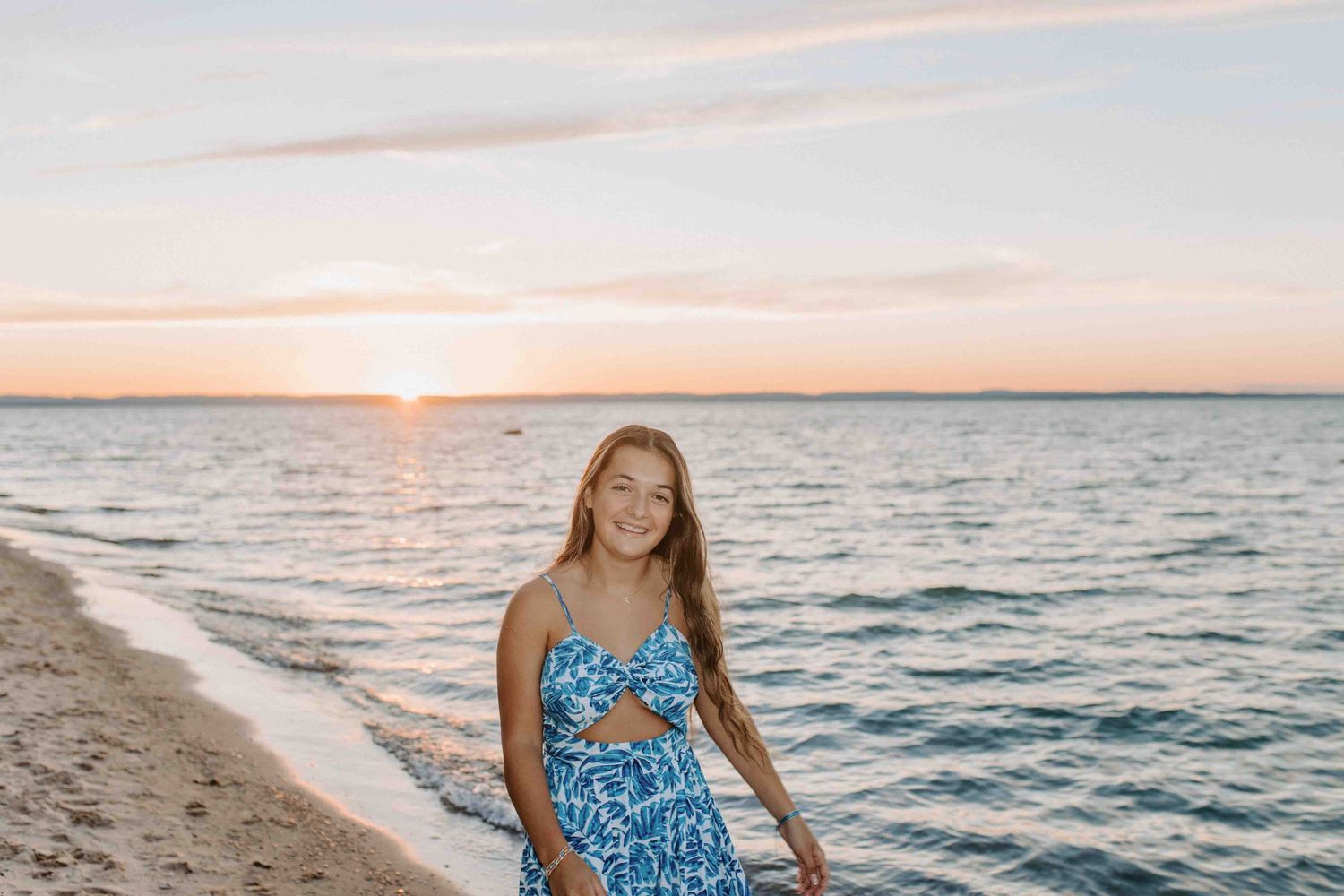 Person in a blue patterned sundress stands on sandy beach with sunset over water.