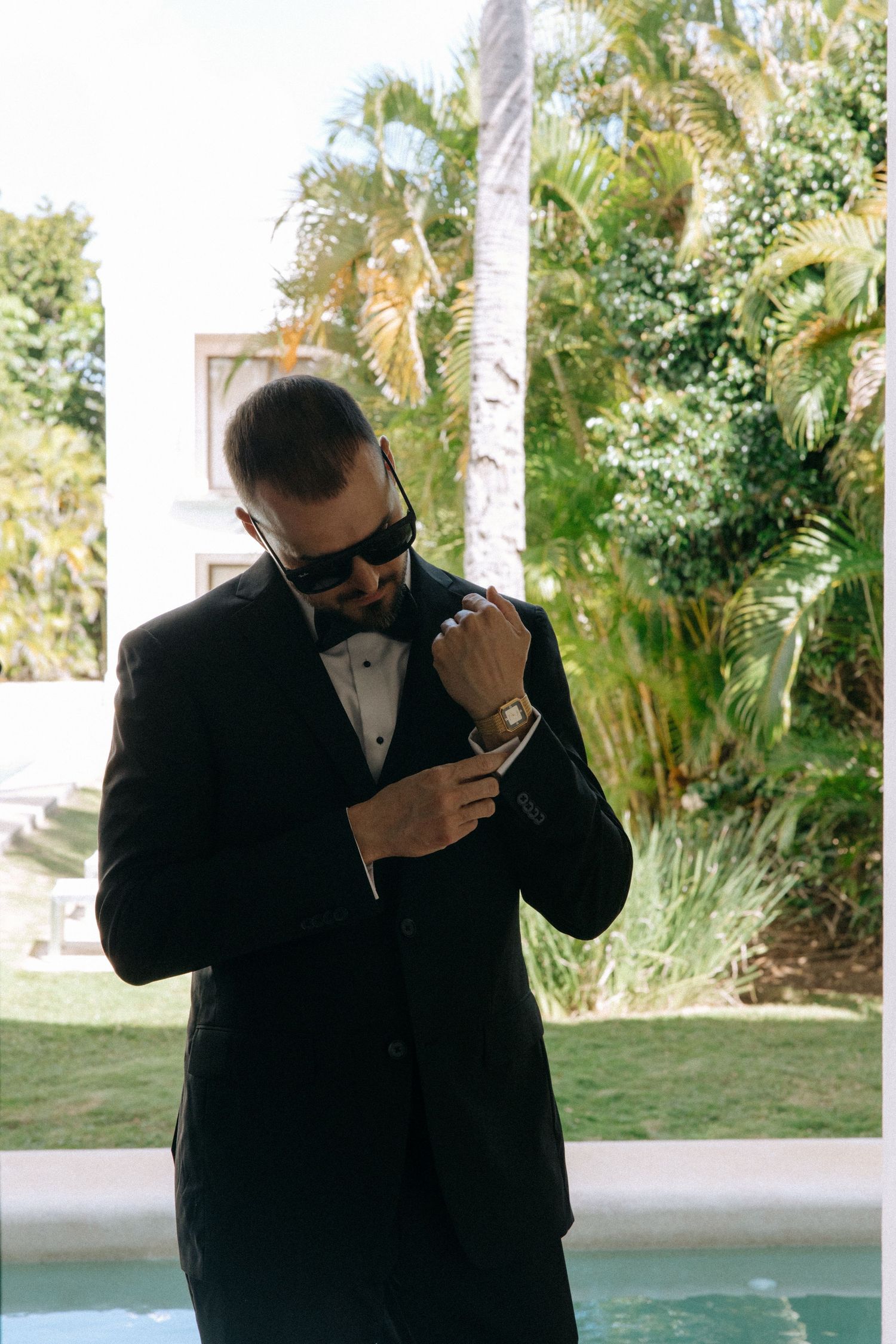 Men in formal black suits embrace outdoors with palm trees in the background.