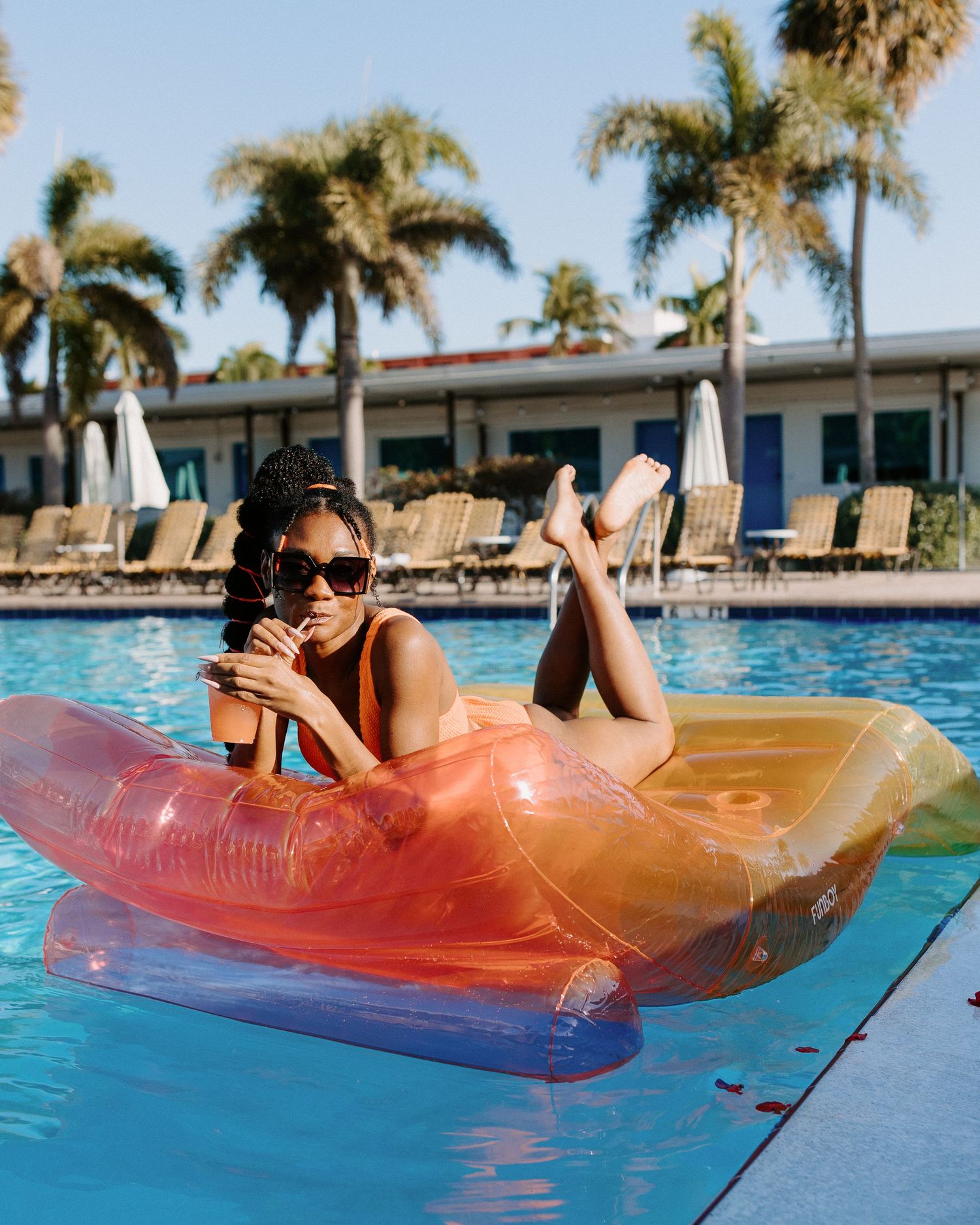 Someone relaxes on a colorful inflatable float in a swimming pool surrounded by palm trees and lounge chairs.
