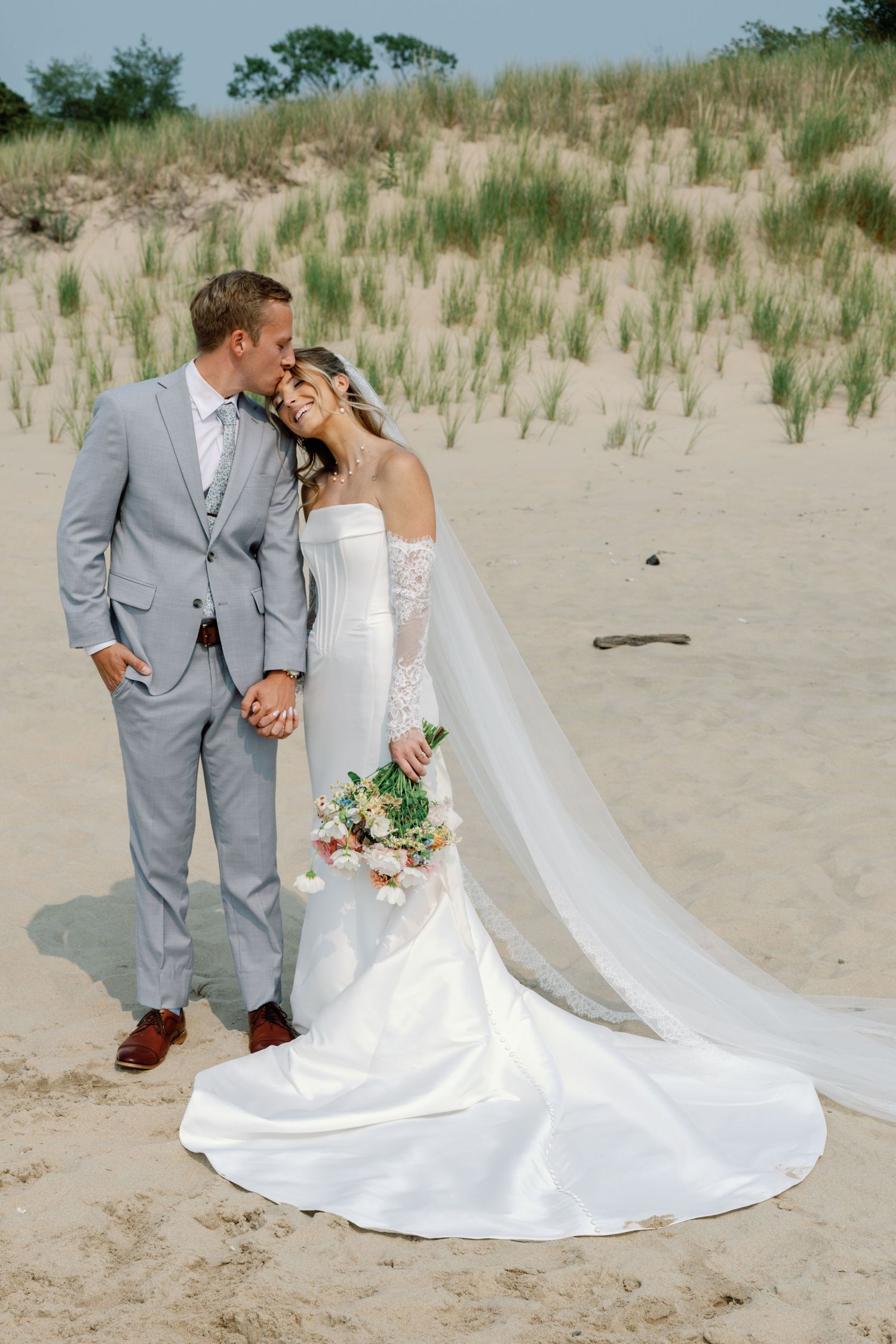 Wedding couple poses together on sandy dunes with flowing white dress and veil.
