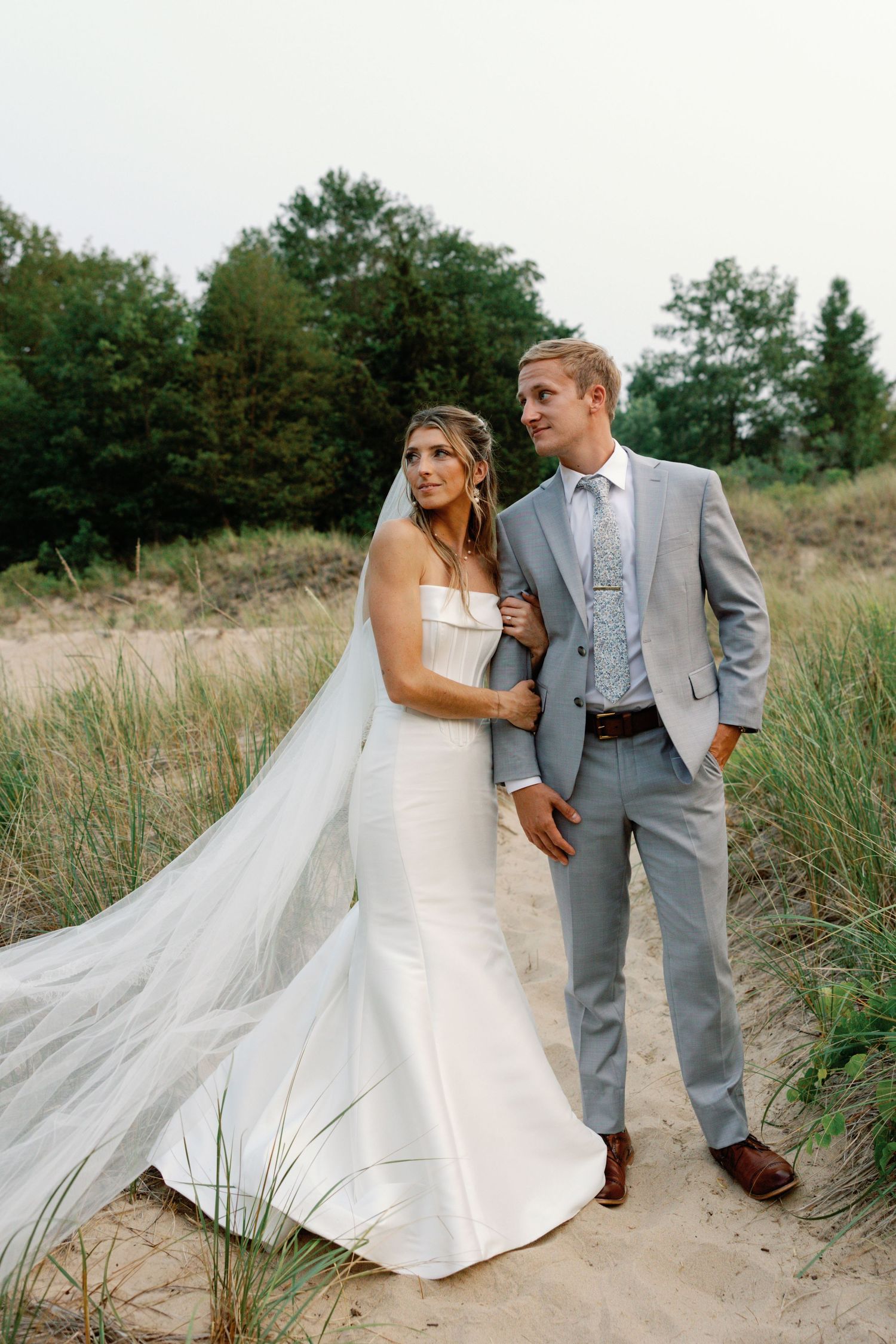 Wedding couple walking along sandy beach path with long veil flowing in wind.