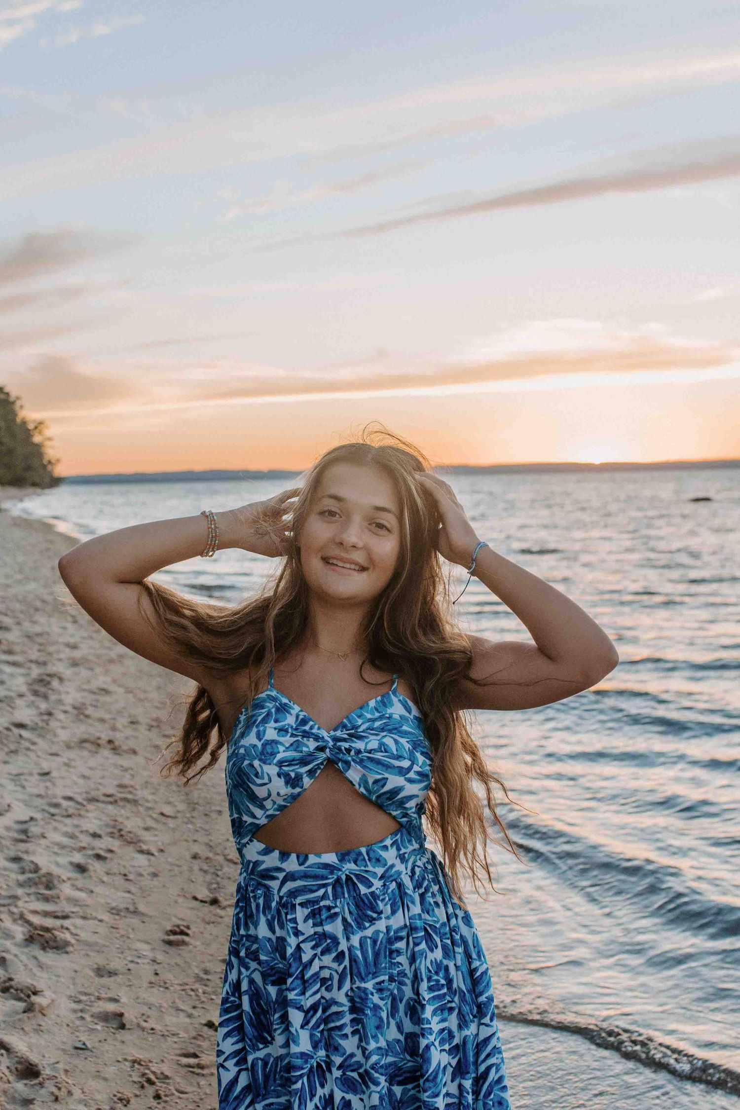 Person in a blue patterned dress poses joyfully on a beach during golden hour sunset.