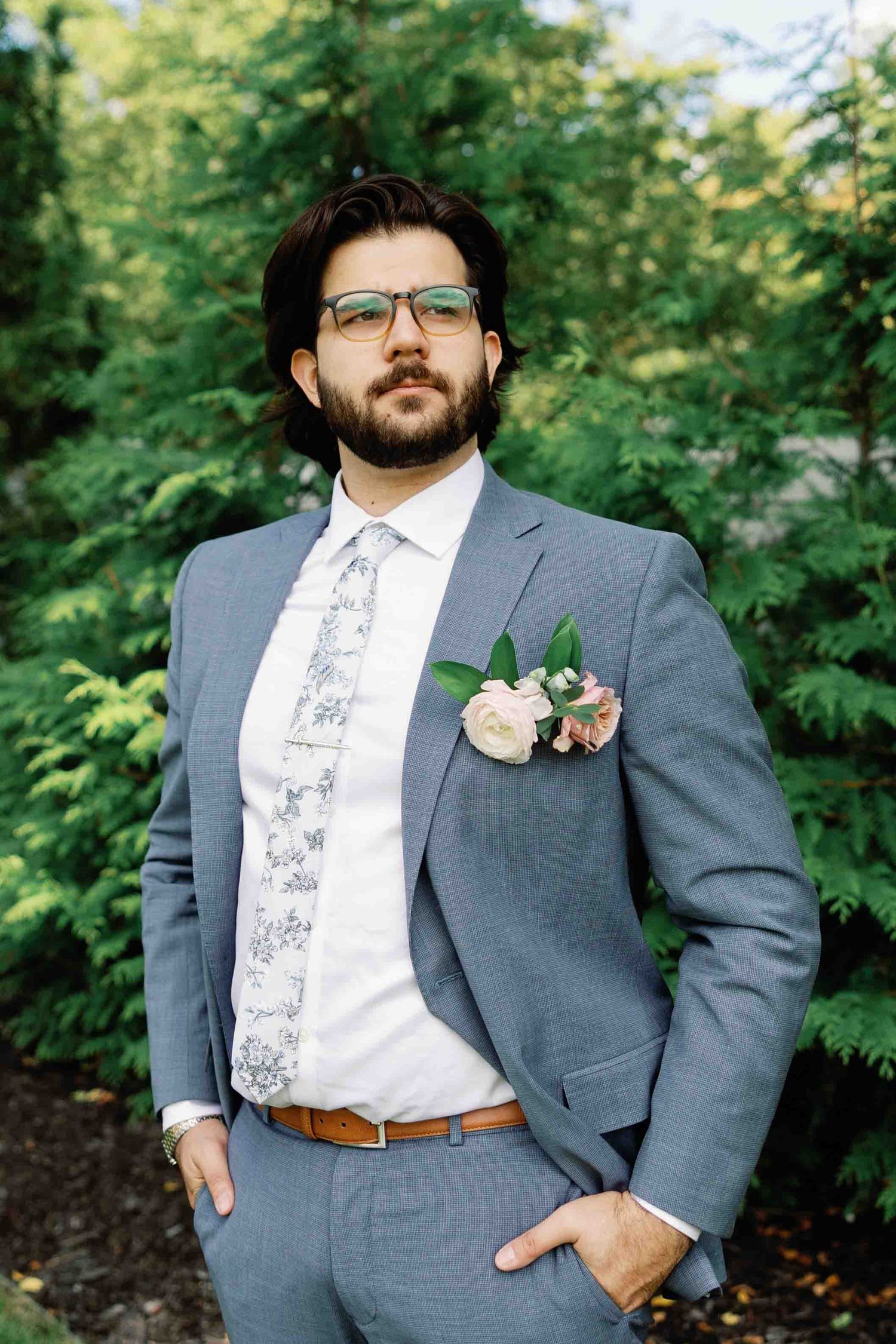 A person in a gray suit with floral boutonniere poses against a backdrop of green trees.