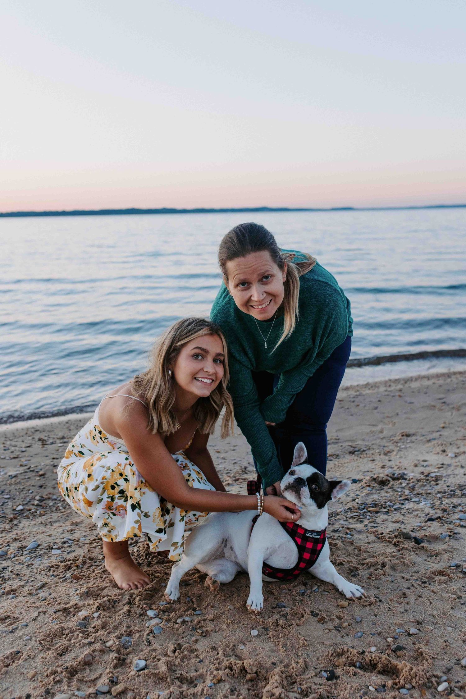 Two people kneel beside a black and white dog on a beach at sunset.