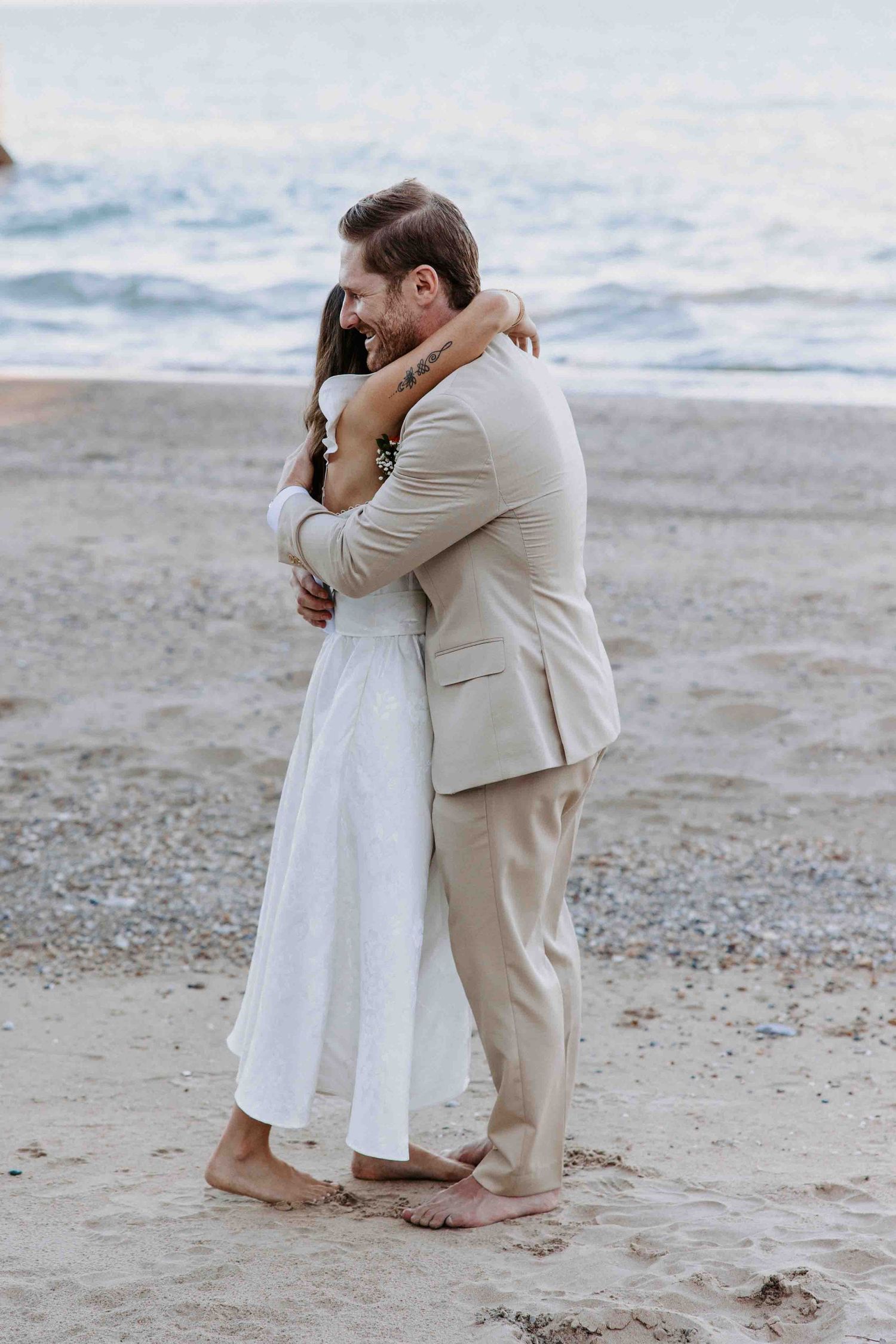 A romantic beach photoshoot featuring a couple embracing in white and beige attire on the sandy shore.