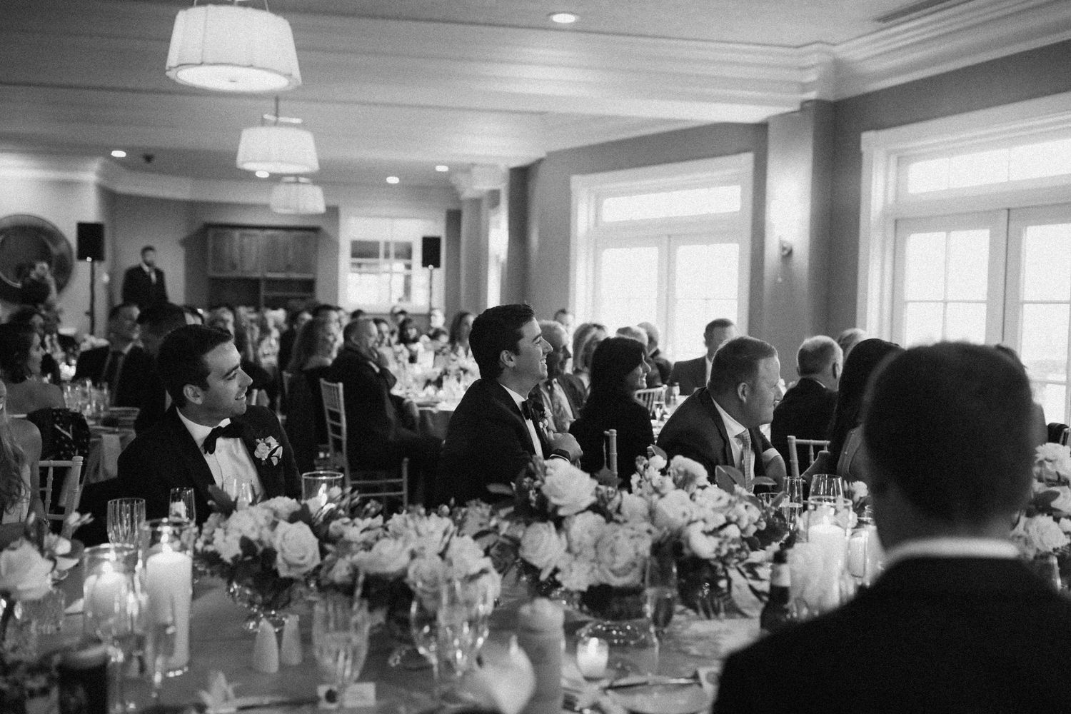 Wedding reception guests gathered in a formal dining room with elegant floral centerpieces and chandeliers.