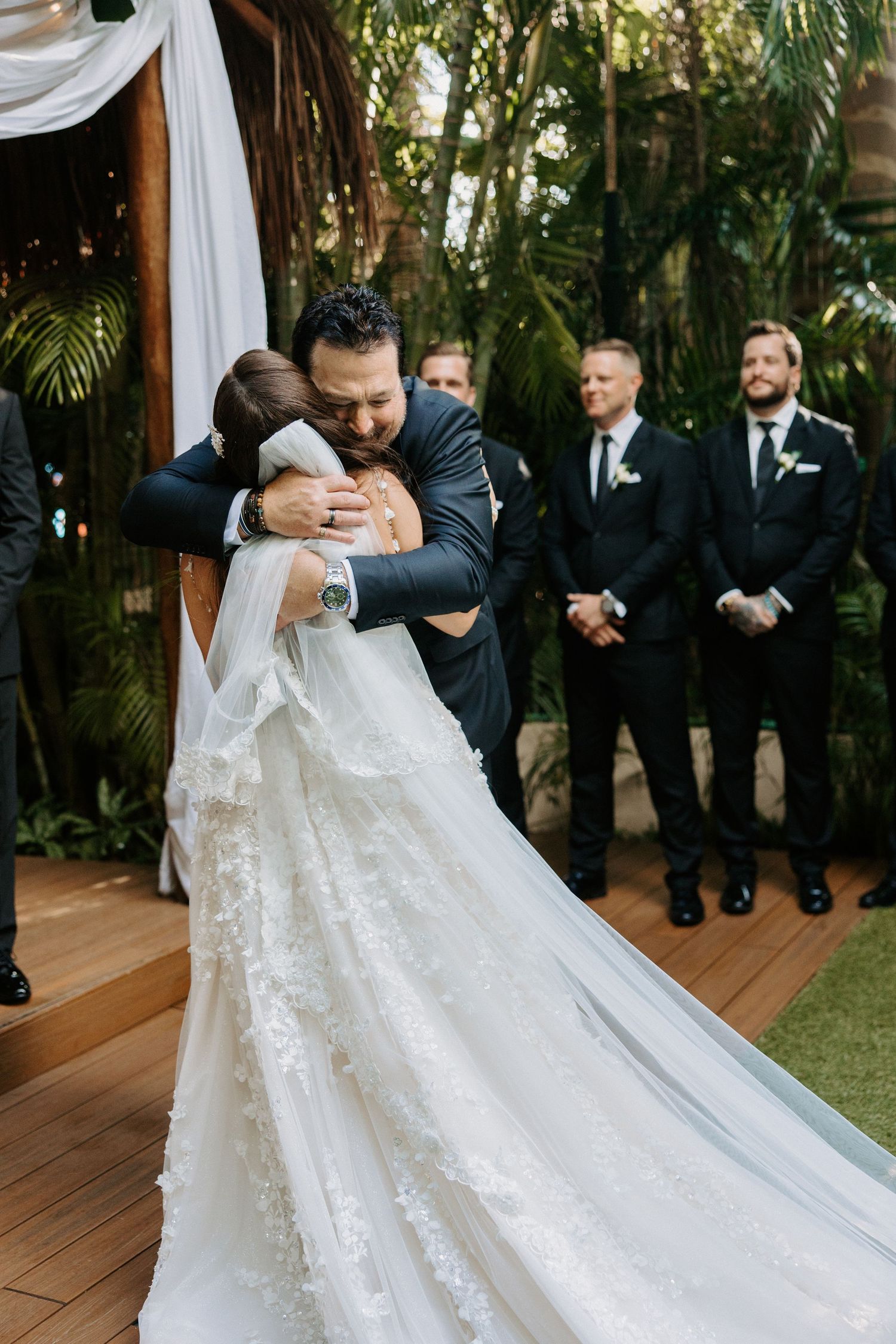 Emotional embrace during outdoor garden wedding ceremony with groomsmen standing on wooden deck surrounded by greenery.