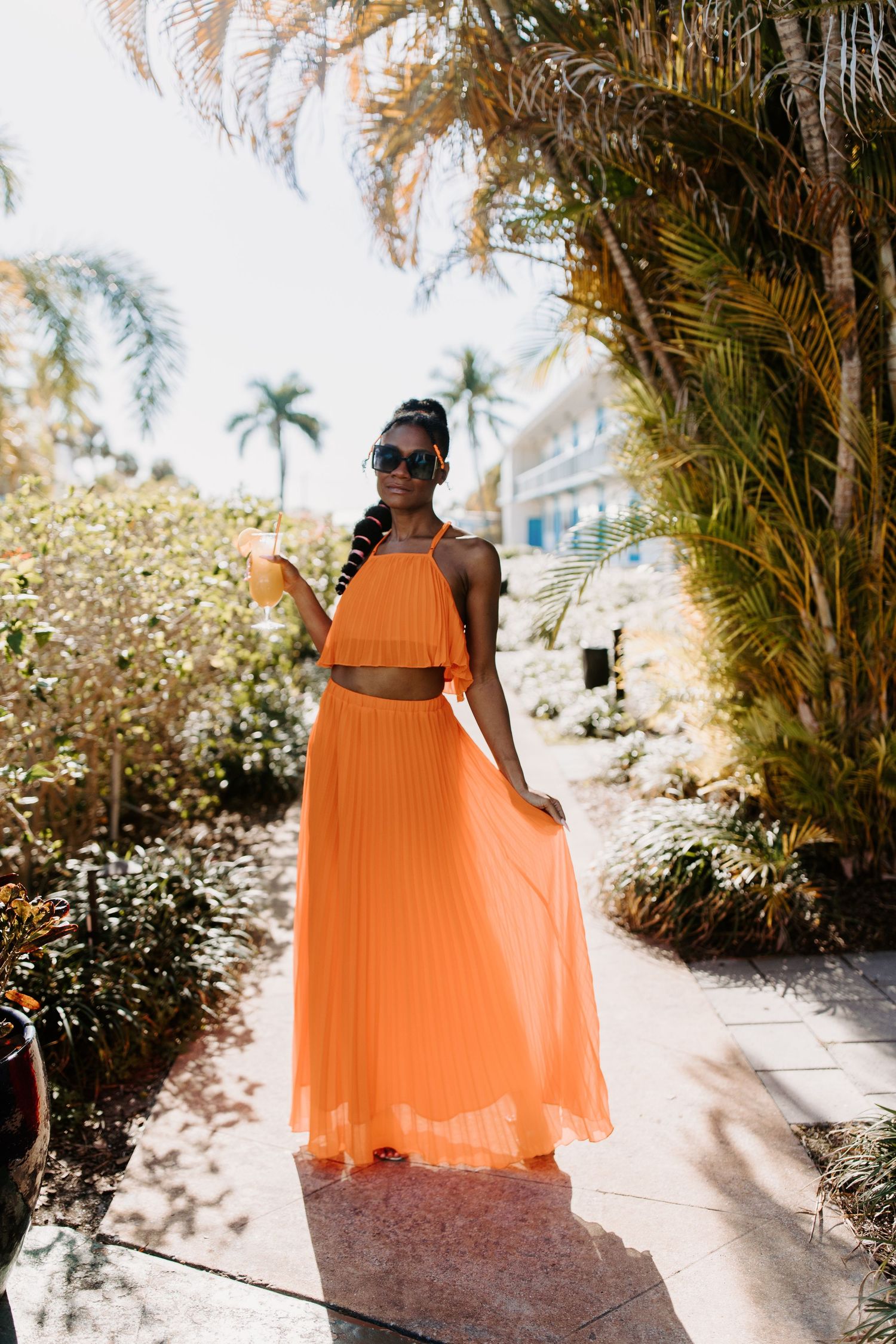 Woman in flowing orange pleated maxi dress poses on tropical garden path with palm trees and sunlit backdrop.