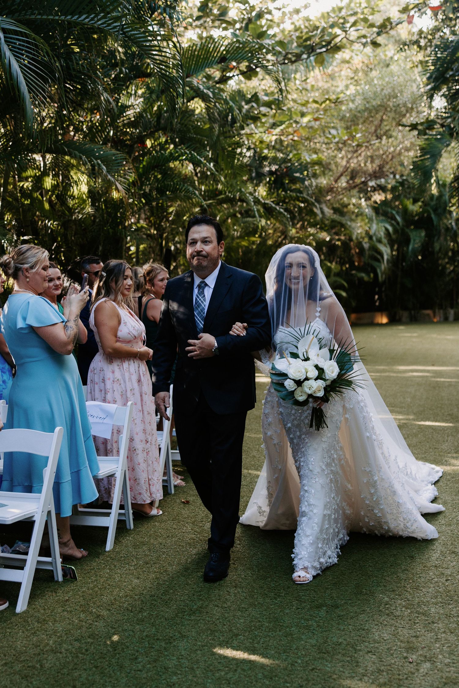 A wedding ceremony takes place outdoors with palm trees and greenery, as attendees watch from white folding chairs.